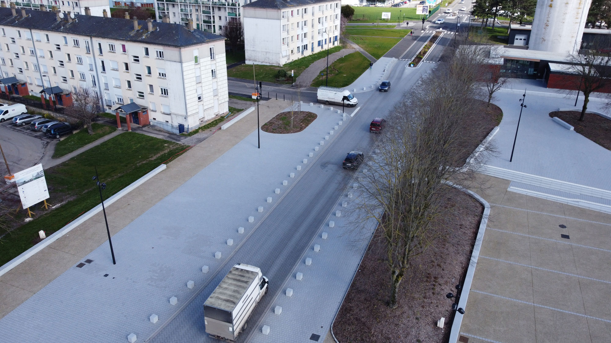 Vue aérienne d'une rue de la ville avec une circulation légère, notamment un camion et plusieurs voitures. Des bâtiments sont visibles à gauche et une allée bordée d'arbres se trouve à droite. La route est bordée de zones pavées et herbeuses.