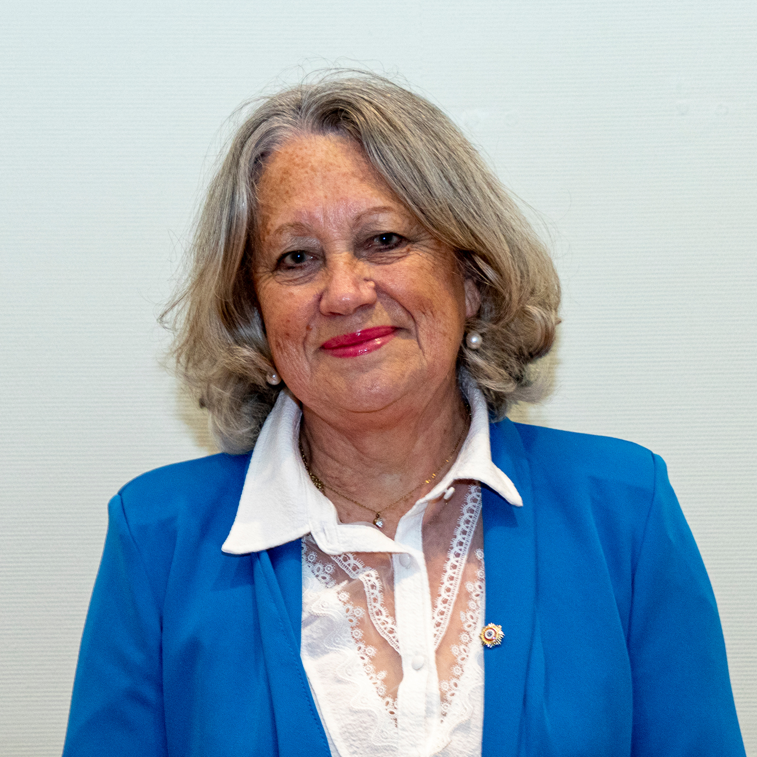 Une femme âgée aux cheveux gris sourit. Elle porte un blazer bleu sur un chemisier blanc, accessoirisé de boucles d'oreilles en perles, d'une petite broche et d'un collier. L'arrière-plan est uni et de couleur claire.