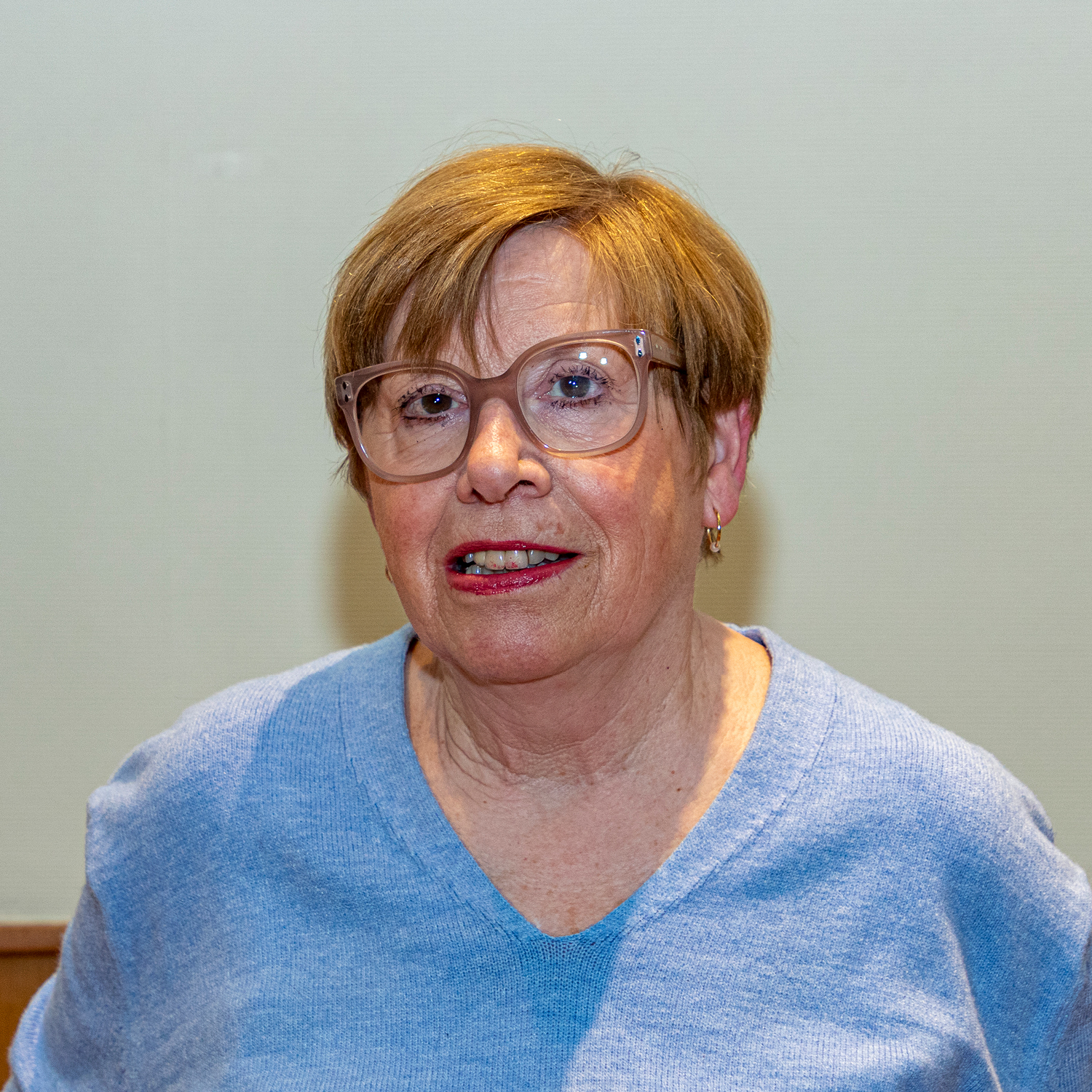 Une femme aux cheveux bruns courts et aux lunettes sourit. Elle porte un pull bleu clair et des boucles d'oreilles dorées. L'arrière-plan est uni et de couleur neutre.