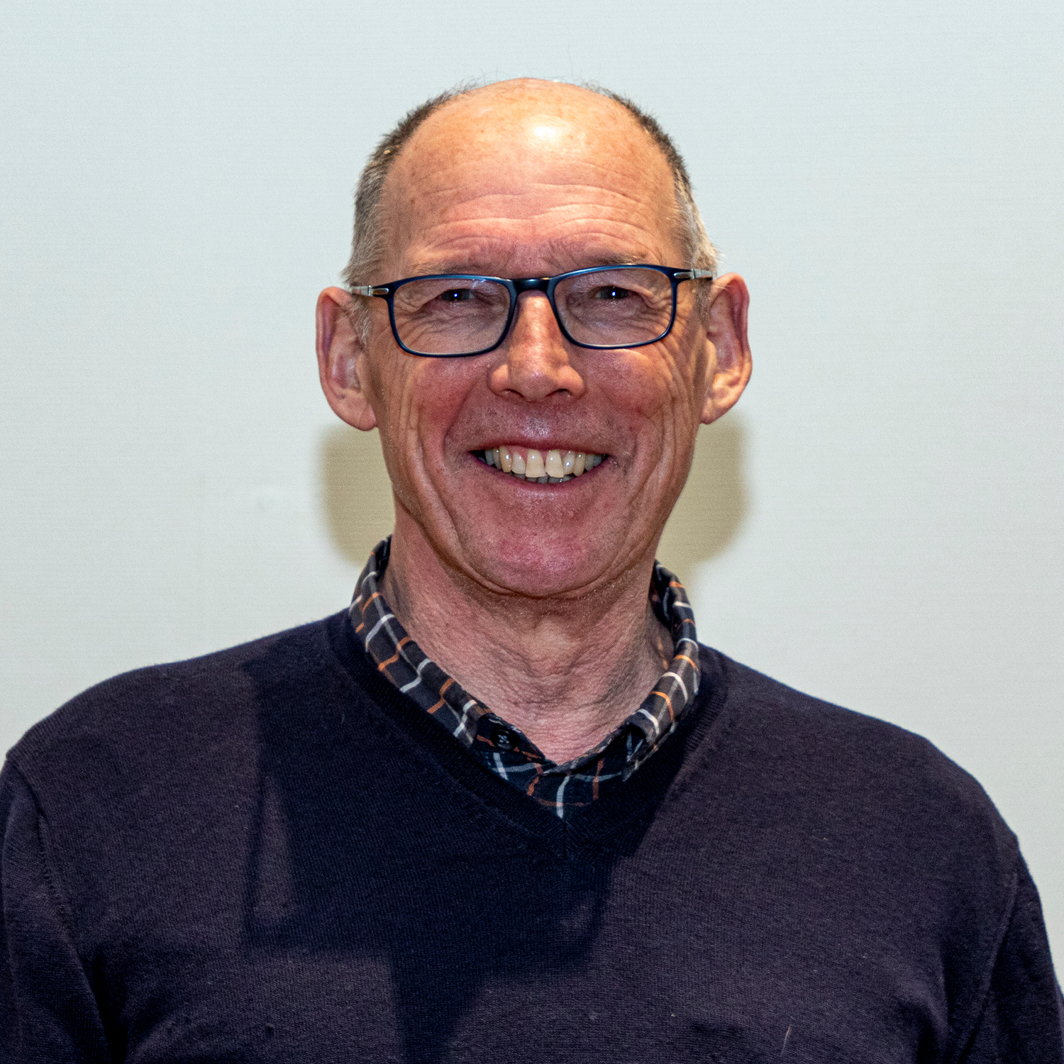 Un homme âgé souriant, portant des lunettes et les cheveux courts, porte un pull foncé sur une chemise à carreaux. Il se tient debout devant un fond blanc uni.