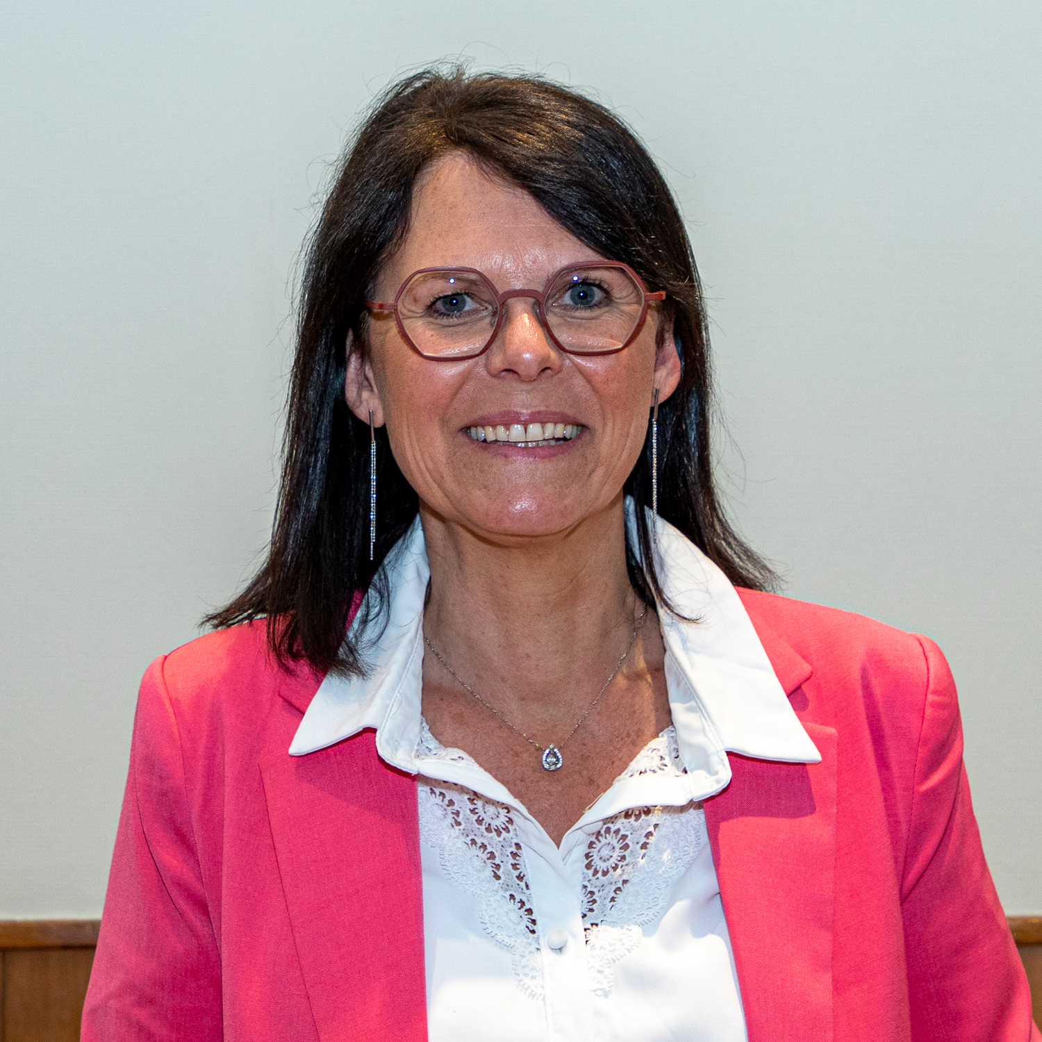 Une femme aux cheveux noirs mi-longs et portant des lunettes sourit tout en portant un blazer rose sur un chemisier blanc. Elle se tient debout sur un fond uni et sa tenue comporte des détails en dentelle.