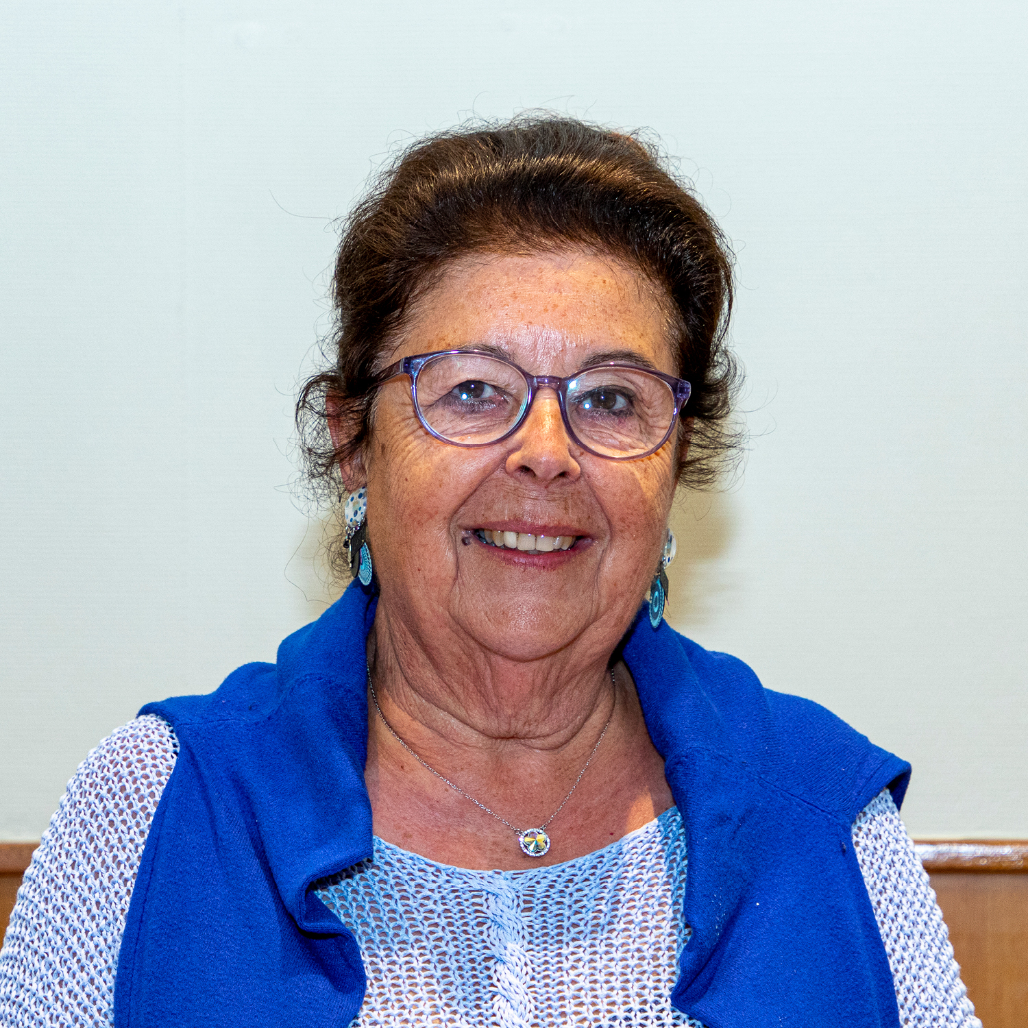 Une femme âgée portant des lunettes sourit à la caméra. Elle porte un gilet bleu sur un haut à motifs blanc et bleu et porte des boucles d'oreilles et un collier assortis. L'arrière-plan est uni et de couleur claire.