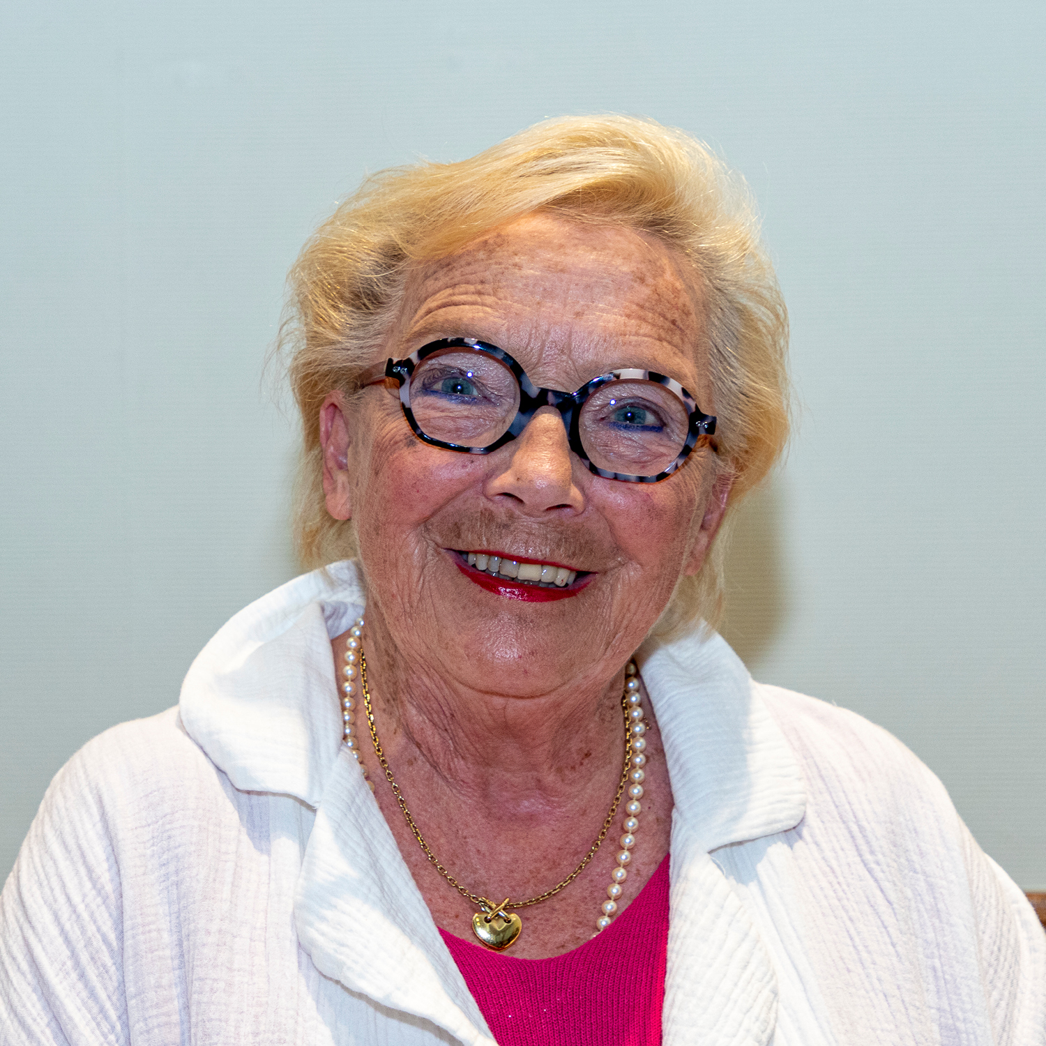 Une femme âgée aux cheveux blonds courts et aux lunettes sourit chaleureusement. Elle porte un chemisier blanc à col sur un haut rose et un collier de perles avec un pendentif en or. L'arrière-plan est un mur uni et clair.