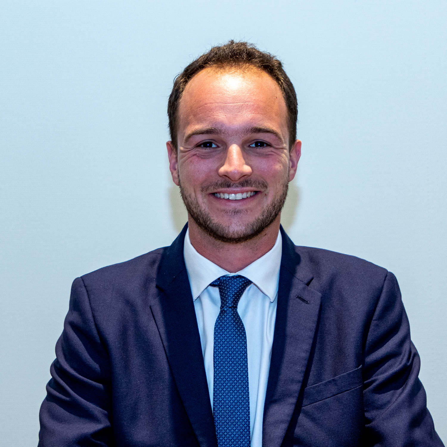 Un homme souriant, vêtu d'un costume bleu foncé, d'une chemise blanche et d'une cravate bleue, se tient debout sur un fond uni de couleur claire.