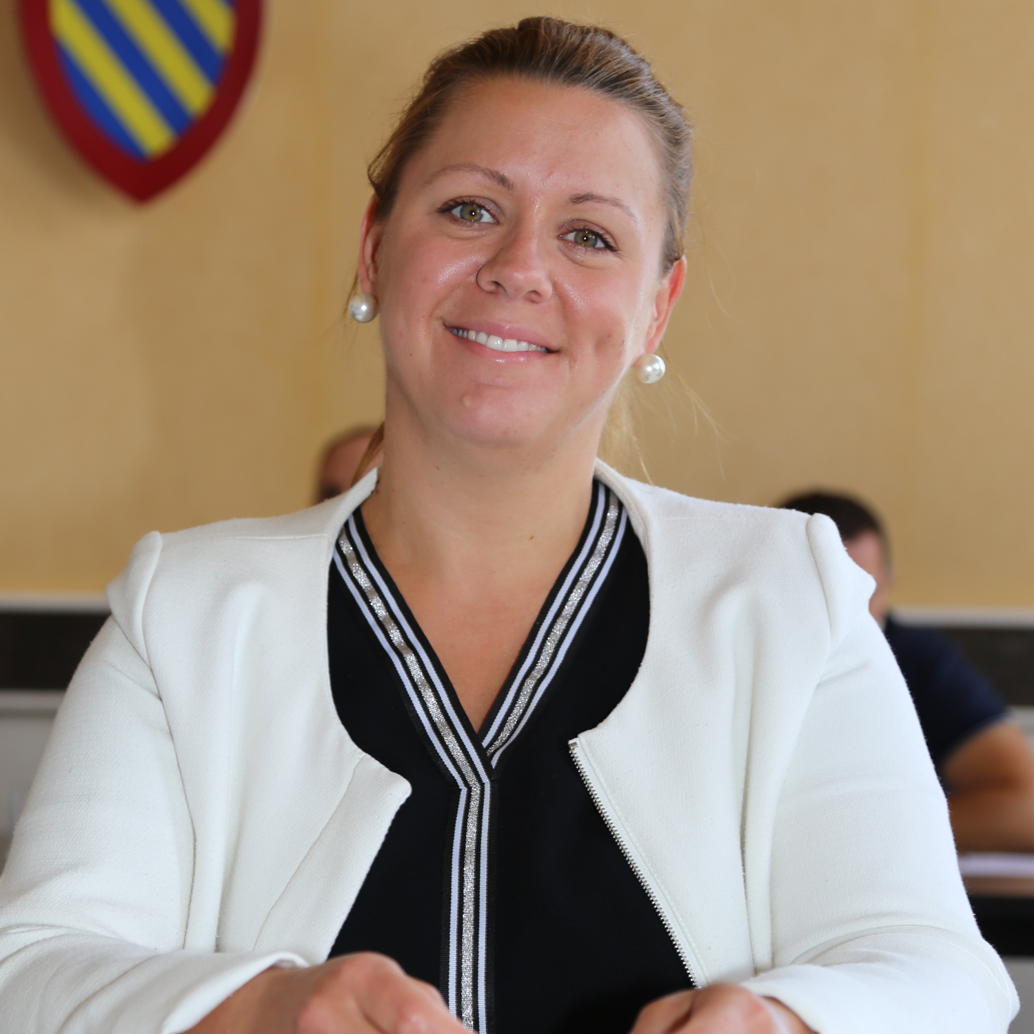 Une femme portant un blazer blanc et un haut noir sourit à la caméra. Elle est assise à l'intérieur avec des personnes floues et un bouclier coloré en arrière-plan.