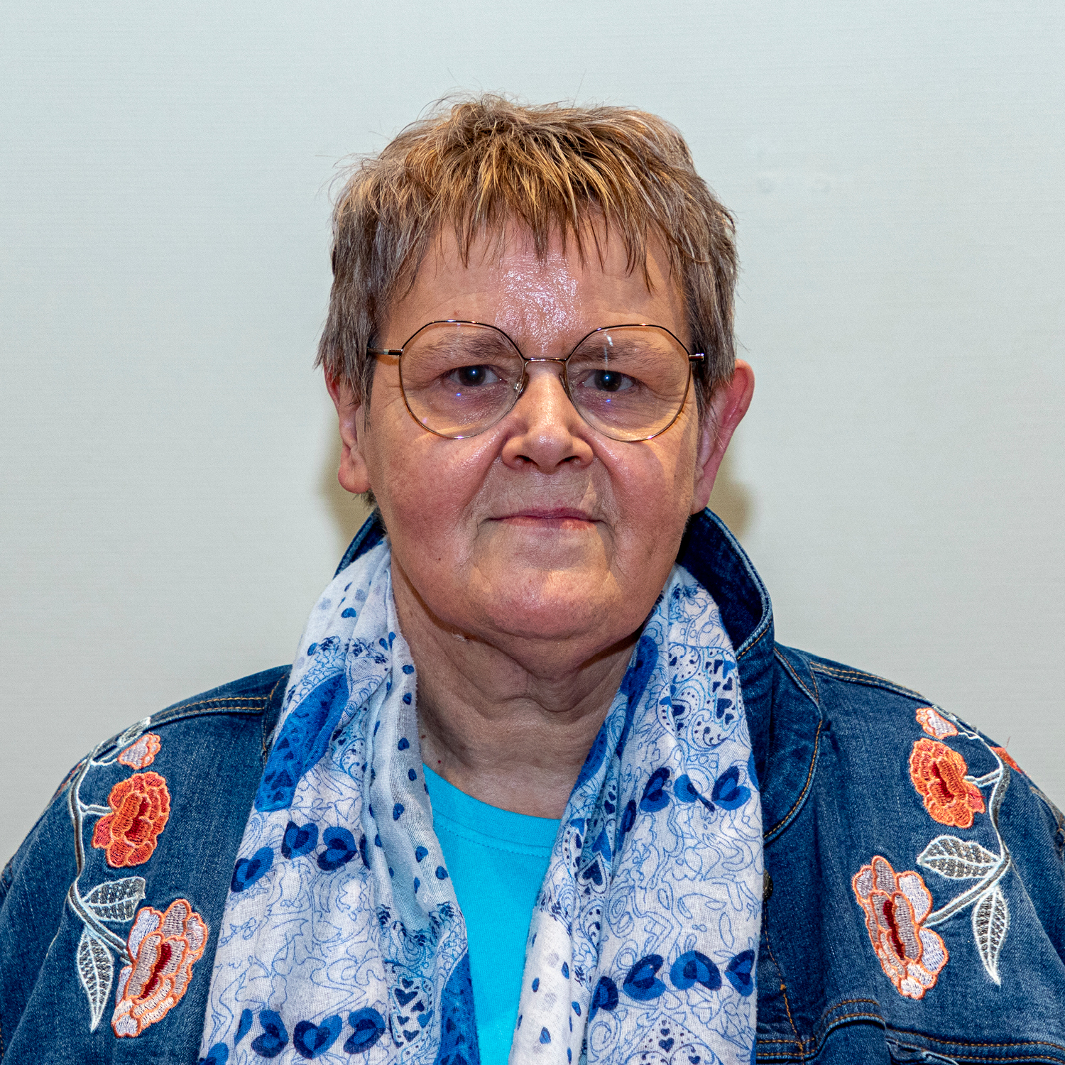 Un adulte âgé portant des lunettes, une veste en jean avec des fleurs brodées et un foulard à motifs bleus se tient debout sur un fond uni. Il a les cheveux courts et une expression neutre.