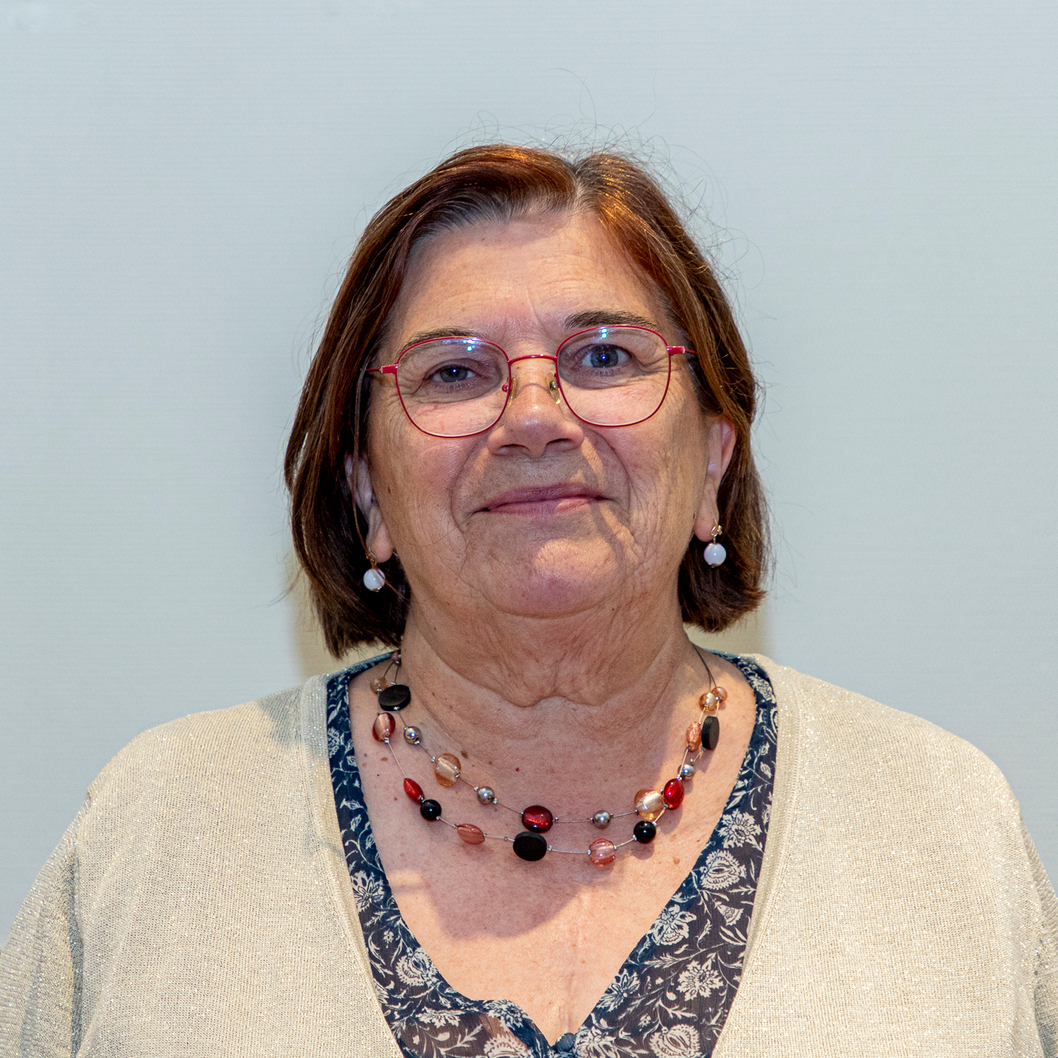 Une femme aux cheveux bruns courts, portant des lunettes et un haut fleuri sous un cardigan beige. Elle porte également un collier de perles. Le fond est d'une couleur claire unie.