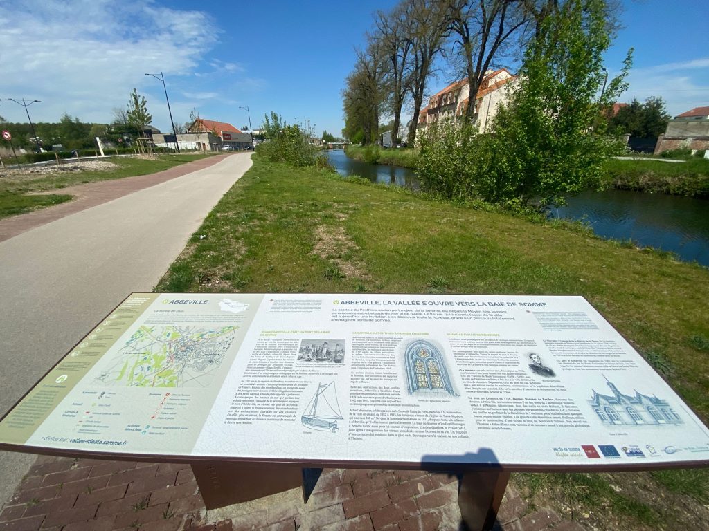 Un panneau d'information panoramique en plein air se dresse le long d'un chemin pavé, avec une rivière et des arbres en arrière-plan. Le panneau contient du texte et des images sur Abbeville, ainsi qu'une carte et des illustrations architecturales. Le ciel est clair et ensoleillé.