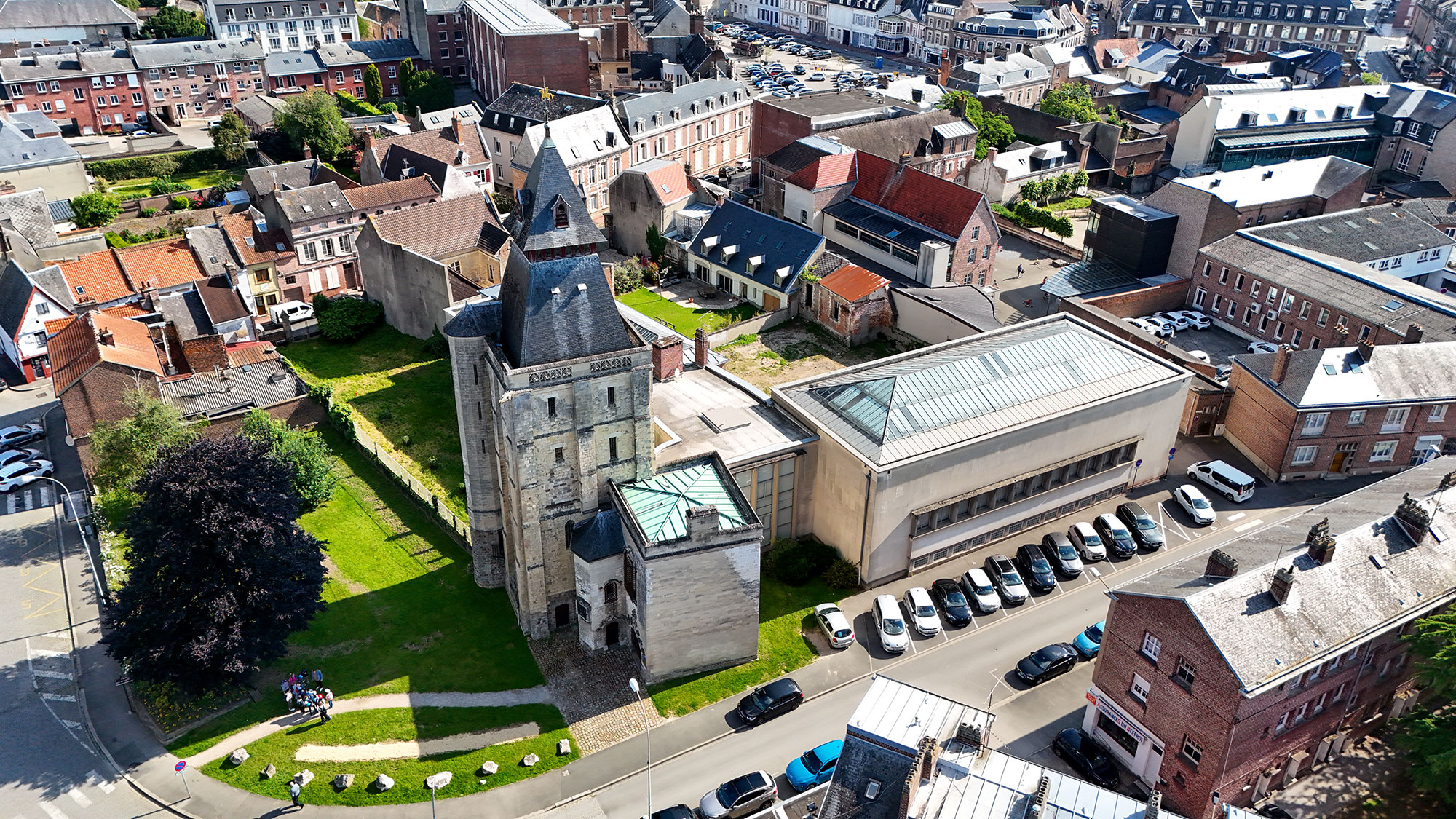 Vue aérienne d'une vieille église en pierre avec un haut clocher, entourée de bâtiments de banlieue et de voitures garées. Un sentier traverse la zone herbeuse autour de l'église, avec des arbres et des gens qui marchent à proximité. Le quartier est densément peuplé et urbain.