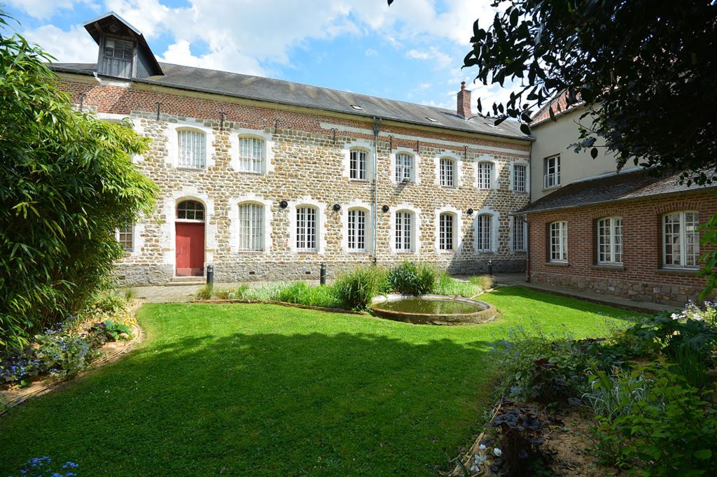 Un bâtiment historique en brique et en pierre avec des fenêtres cintrées et une porte rouge, entouré d'un jardin verdoyant et luxuriant. Un petit étang circulaire se trouve au centre de l'herbe, avec des plantes feuillues et des fleurs le long des bordures.