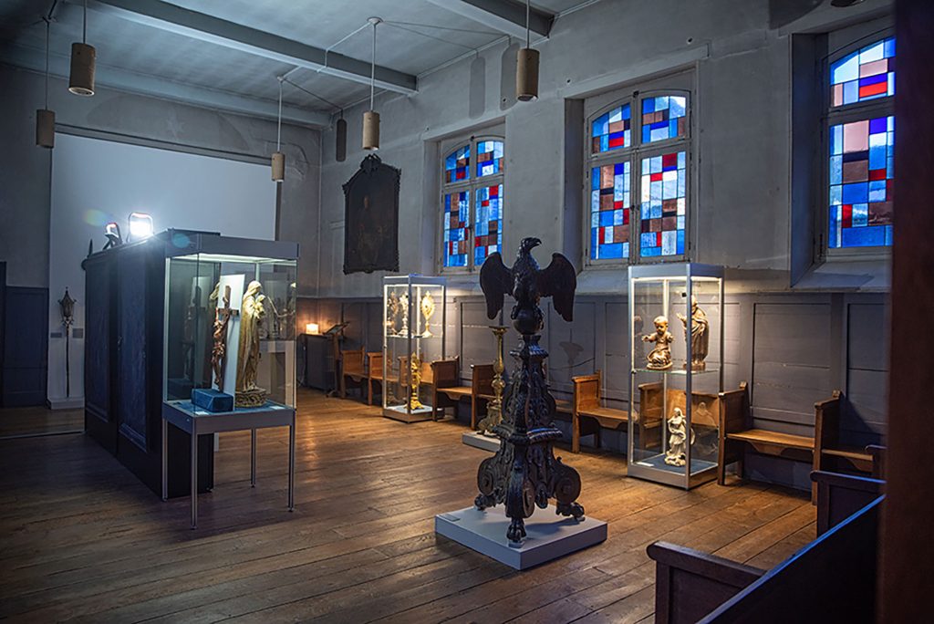 Une salle de musée faiblement éclairée présentant des statues dans des vitrines en verre. La salle est dotée de parquet et de bancs en bois, ainsi que de vitraux colorés laissant entrer une lumière douce. Une grande sculpture d'aigle trône au centre.