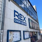 Vue extérieure du Centre Culturel REX, un centre culturel avec une grande enseigne sur un mur blanc. Sous l'enseigne se trouvent des vitrines contenant diverses affiches et informations. Le bâtiment est doté de grandes fenêtres en verre et le ciel est clair et bleu.