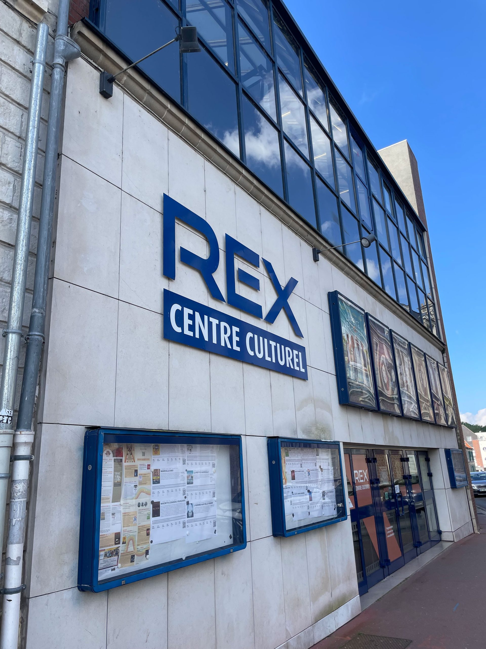 Vue extérieure du Centre Culturel REX, un centre culturel avec une grande enseigne sur un mur blanc. Sous l'enseigne se trouvent des vitrines contenant diverses affiches et informations. Le bâtiment est doté de grandes fenêtres en verre et le ciel est clair et bleu.