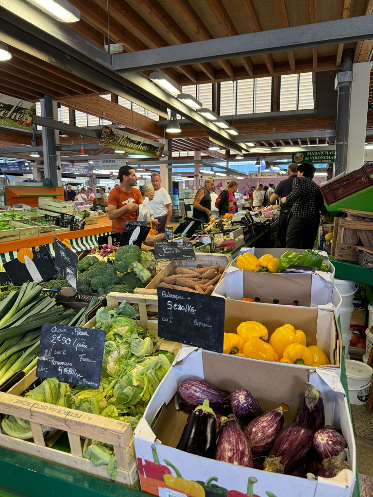 Un marché couvert animé avec divers légumes frais exposés, notamment des aubergines, des poivrons et du brocoli. Les clients parcourent les étals sous un plafond en bois, avec des panneaux de prix manuscrits visibles.