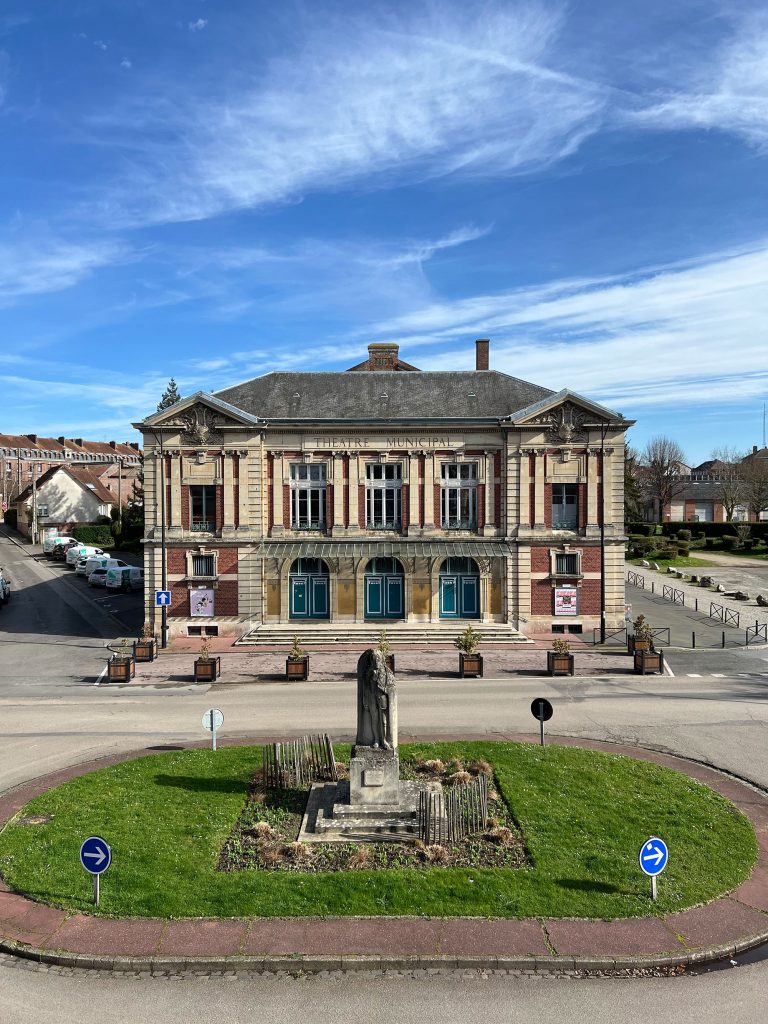 Un bâtiment historique avec une façade décorative se dresse sous un ciel bleu. Devant, un petit rond-point avec une statue sur une île herbeuse est entouré de panneaux de signalisation. Des bâtiments résidentiels sont visibles à l'arrière-plan.