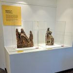 Deux statues religieuses exposées dans des vitrines en verre sur un piédestal blanc dans le cadre d'une exposition intitulée « Univers 11 : Trésors d'art et d'architecture ». L'une des statues représente une scène biblique avec trois personnages, tandis que l'autre représente une autre figure religieuse.