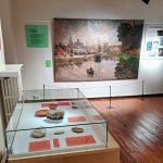 Exposition muséale avec un grand tableau représentant une scène de canal accroché au mur. Au premier plan, une vitrine en verre contient divers objets sur un plancher en bois. Des panneaux d'information sont visibles sur les murs à côté du tableau.