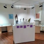 Une exposition de musée avec un piédestal blanc et cinq statues dans une vitrine en verre. La salle est dotée de parquet et de plaques d'information sur les murs. Des projecteurs éclairent l'exposition.