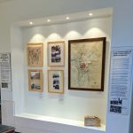 Une exposition de musée présentant des cartes et des photographies historiques exposées sur un mur blanc. La pièce maîtresse est une grande carte encadrée, entourée de photos encadrées plus petites et de panneaux d'information textuelle. Une brique repose sur une petite étagère en dessous.