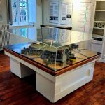 Une maquette 3D détaillée d'une ville historique est posée sur une table en bois dans une salle de musée. La maquette est enfermée dans une vitrine transparente. La salle est dotée de parquet, de panneaux d'information sur les murs et d'une fenêtre avec volets.