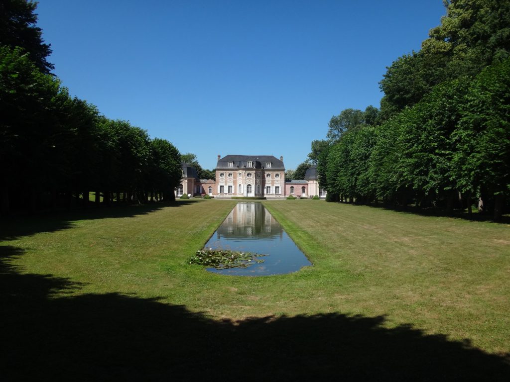 Un grand bâtiment à la façade symétrique se dresse au bout d'une longue pelouse bien entretenue bordée d'arbres. Un étroit bassin réfléchissant traverse la zone herbeuse, guidant le regard vers la structure. Le ciel est clair et bleu.