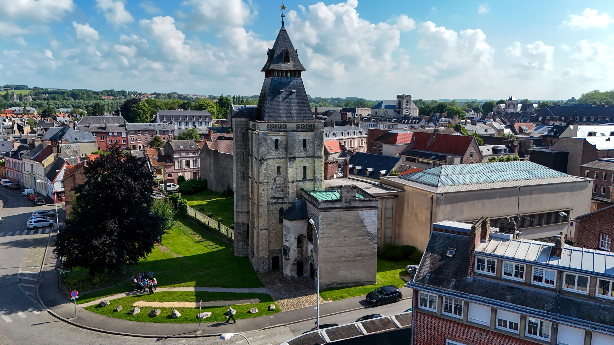 Vue aérienne d'une église historique en pierre avec un haut clocher, entourée d'un mélange de bâtiments anciens et modernes dans une petite ville. Un groupe de personnes est rassemblé près de l'entrée. Le ciel est partiellement nuageux et de la verdure est visible au loin.