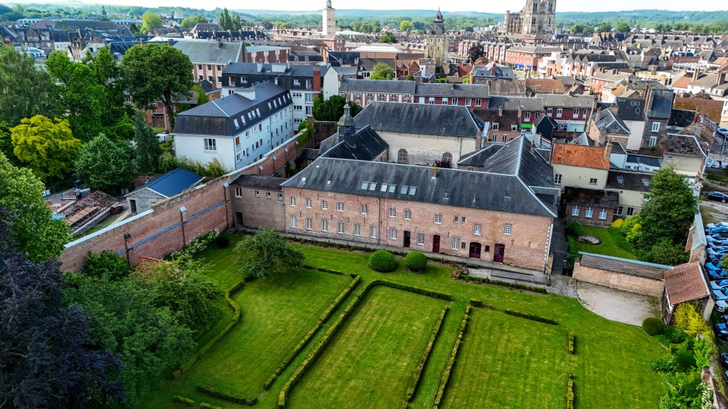 Vue aérienne d'une ville historique européenne avec un grand bâtiment en brique, des jardins bien entretenus et un mélange d'architecture ancienne et moderne. La ville est entourée d'une végétation luxuriante et présente un clocher d'église proéminent en arrière-plan.