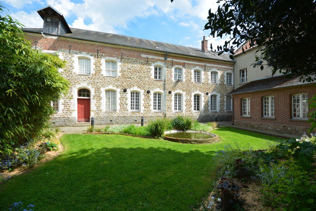 Derrière un jardin bien entretenu se dresse un charmant bâtiment historique en pierre et en brique aux fenêtres encadrées de blanc. Le jardin comprend une pelouse luxuriante et un petit étang circulaire, entouré de diverses plantes et fleurs.