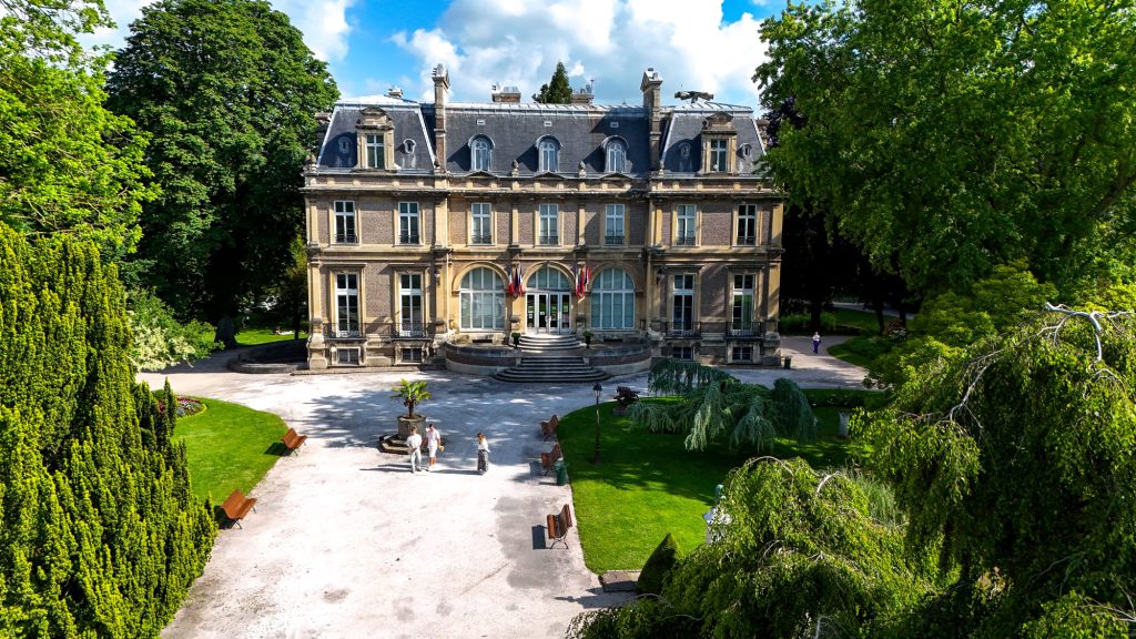 Un grand manoir historique se dresse au milieu d'une végétation luxuriante, avec des jardins symétriques et des bancs bordant le chemin. Le bâtiment présente une architecture ornée avec des fenêtres cintrées et une entrée centrale, soulignée par un ciel bleu clair.