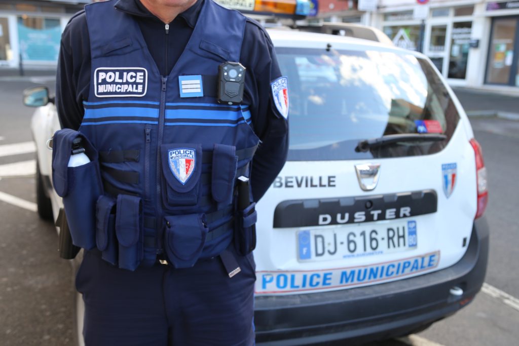 Un policier municipal se tient devant une voiture de police. L'agent porte un uniforme bleu et un gilet avec équipement. La voiture de police derrière porte des plaques d'immatriculation et des insignes visibles. Le décor semble être une rue urbaine.