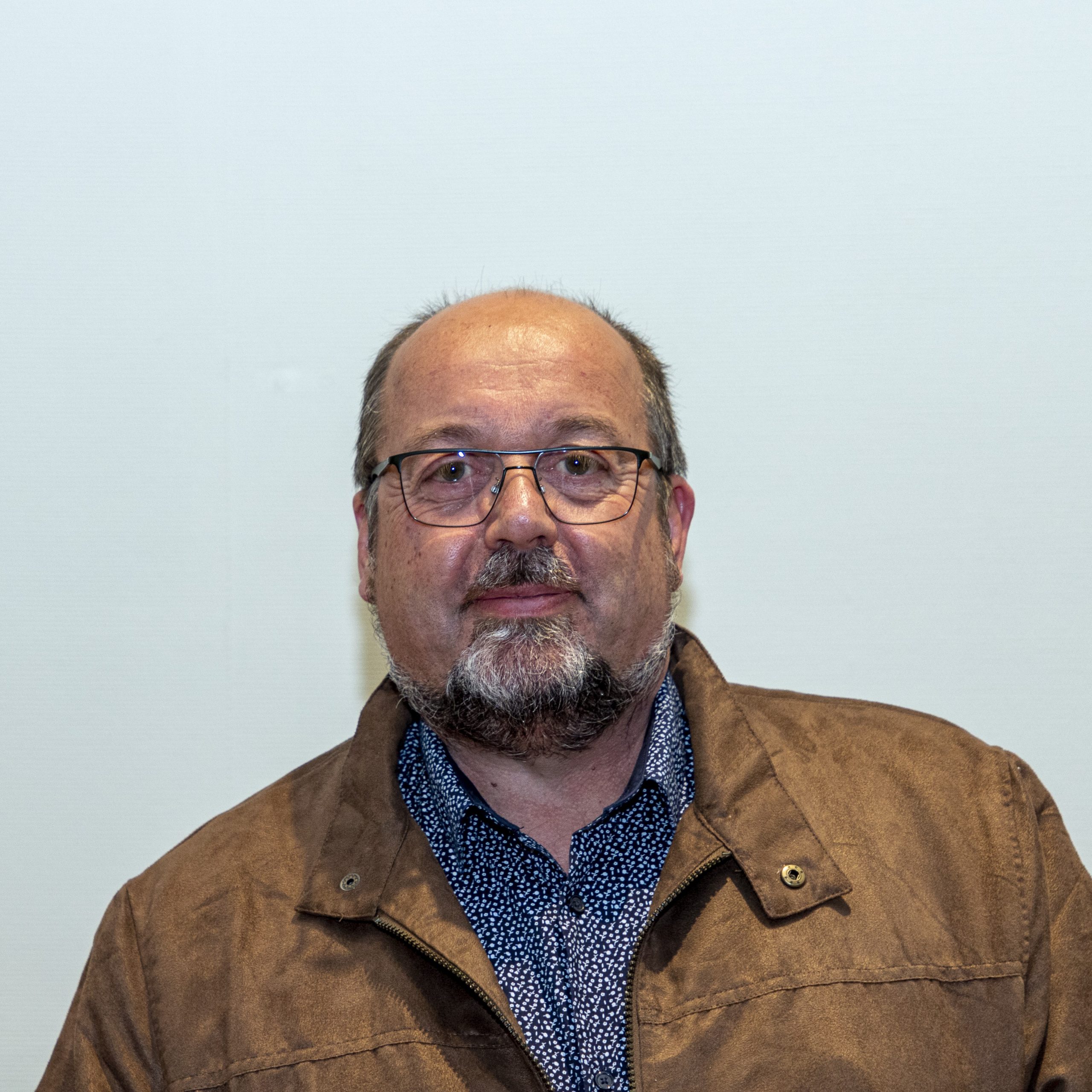 Un homme portant des lunettes et une barbe porte une veste marron sur une chemise bleue à motifs. Il se tient debout sur un fond uni et clair.