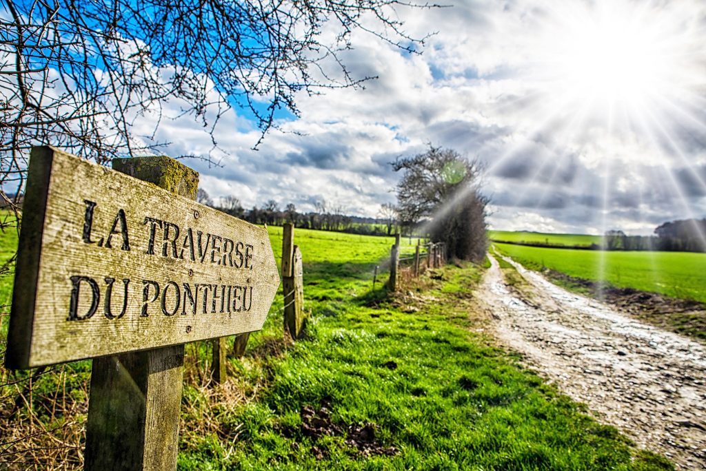 Un panneau en bois indique « La Traverse du Ponthieu » près d'un chemin de terre dans une campagne ensoleillée. Des rayons de soleil brillants brillent à travers le ciel partiellement nuageux au-dessus des champs verts et des arbres nus.