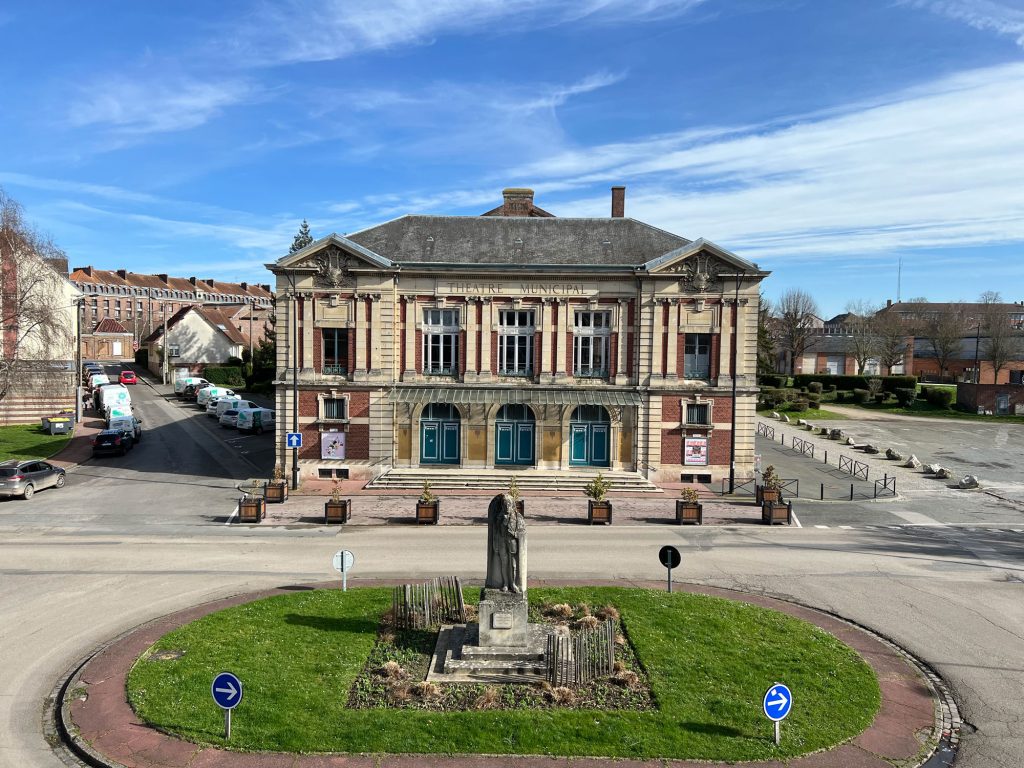 Derrière un petit rond-point agrémenté d'une statue et d'un espace vert se dresse un bâtiment historique en briques aux hautes fenêtres. Le ciel est clair et bleu et plusieurs véhicules stationnés bordent la rue voisine.