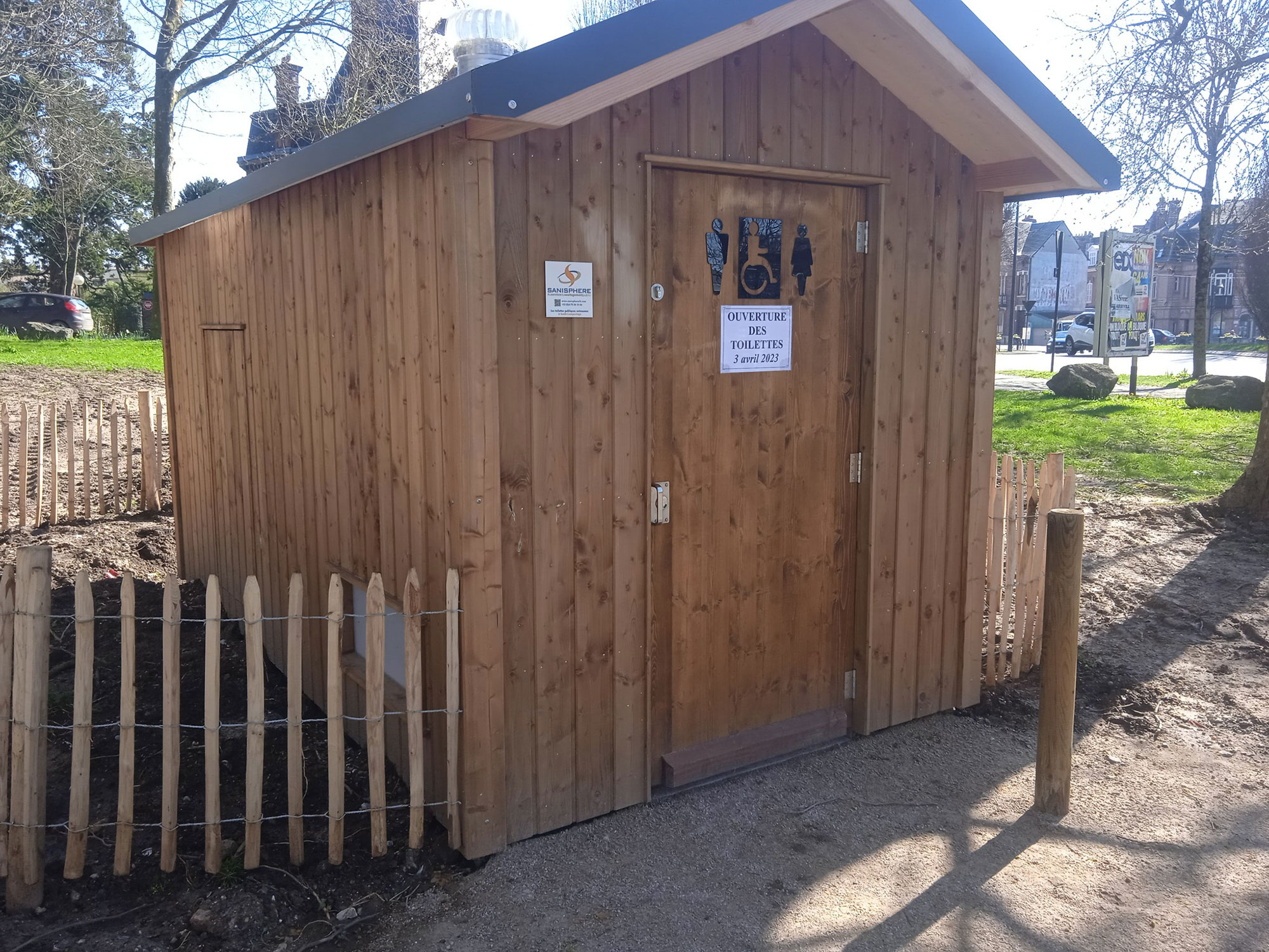 Un petit hangar en bois avec une pancarte sur la porte qui dit "Ouverture tous les jours 24-24". Il est entouré d'une palissade en bois et situé sur un chemin de terre avec des arbres et de l'herbe à proximité.