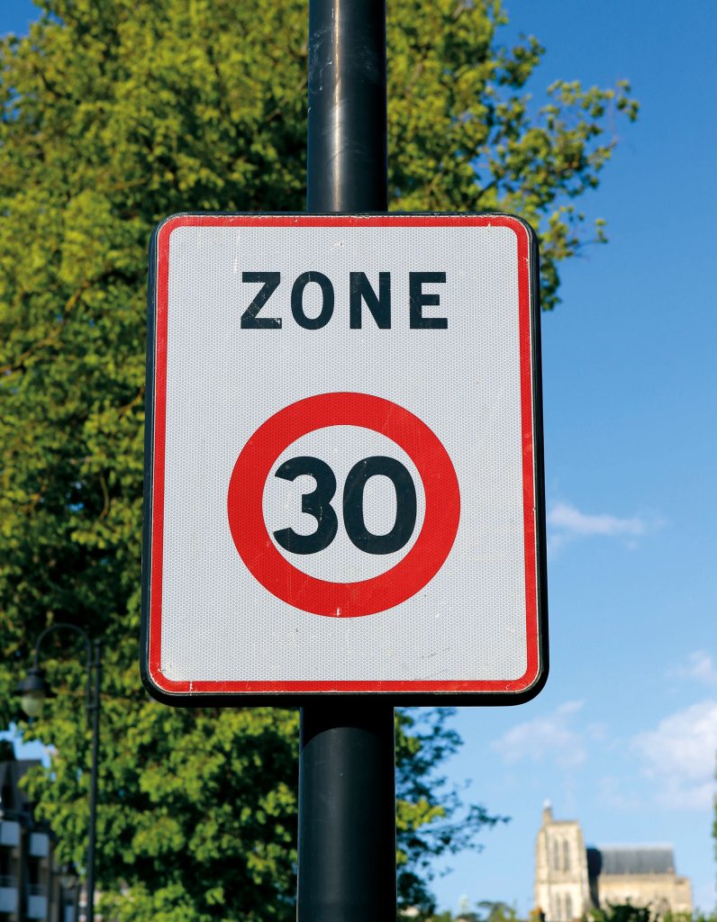 Un panneau de limitation de vitesse indiquant « Zone 30 » avec un cercle rouge autour du numéro 30, monté sur un poteau. Des arbres et un bâtiment sont visibles en arrière-plan sous un ciel bleu clair.