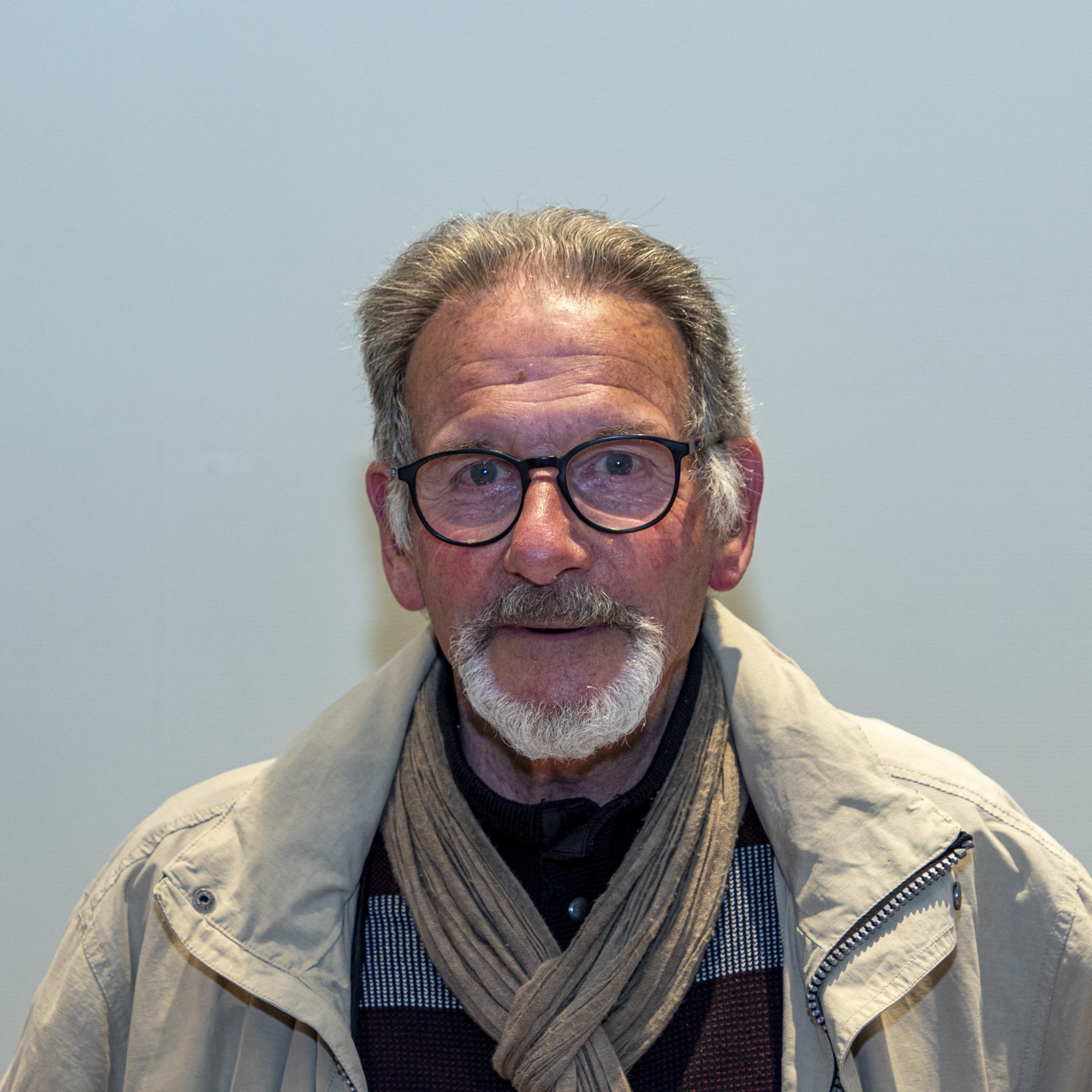 Un homme âgé avec des lunettes et une moustache blanche porte une veste beige et un foulard. Il se tient debout devant un arrière-plan uni, regardant directement la caméra.