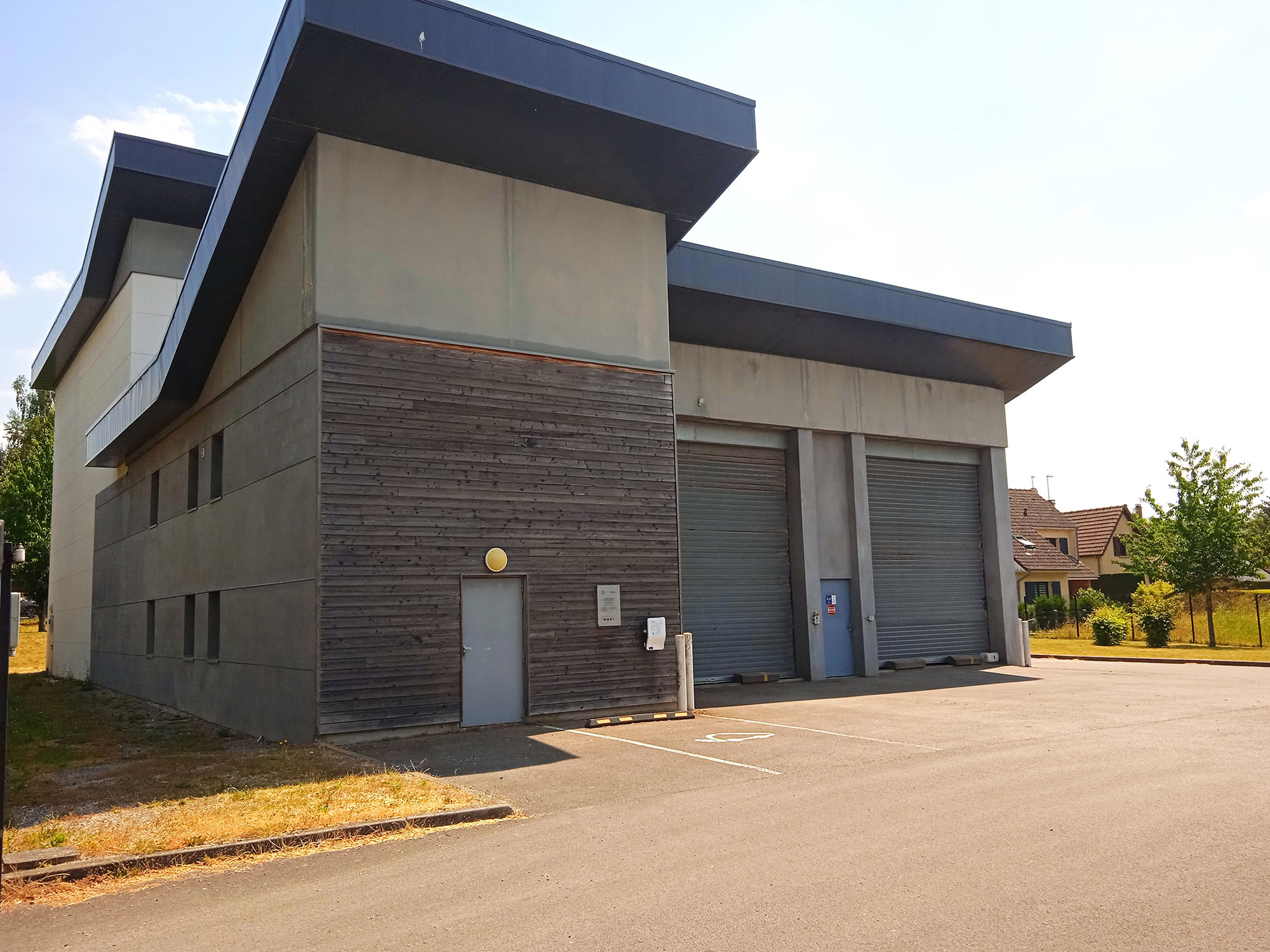 Un bâtiment industriel moderne avec deux grandes portes de garage et une entrée latérale. La façade est un mélange de béton et de panneaux de bois. La zone environnante est pavée d'asphalte et un peu de verdure est visible en arrière-plan.