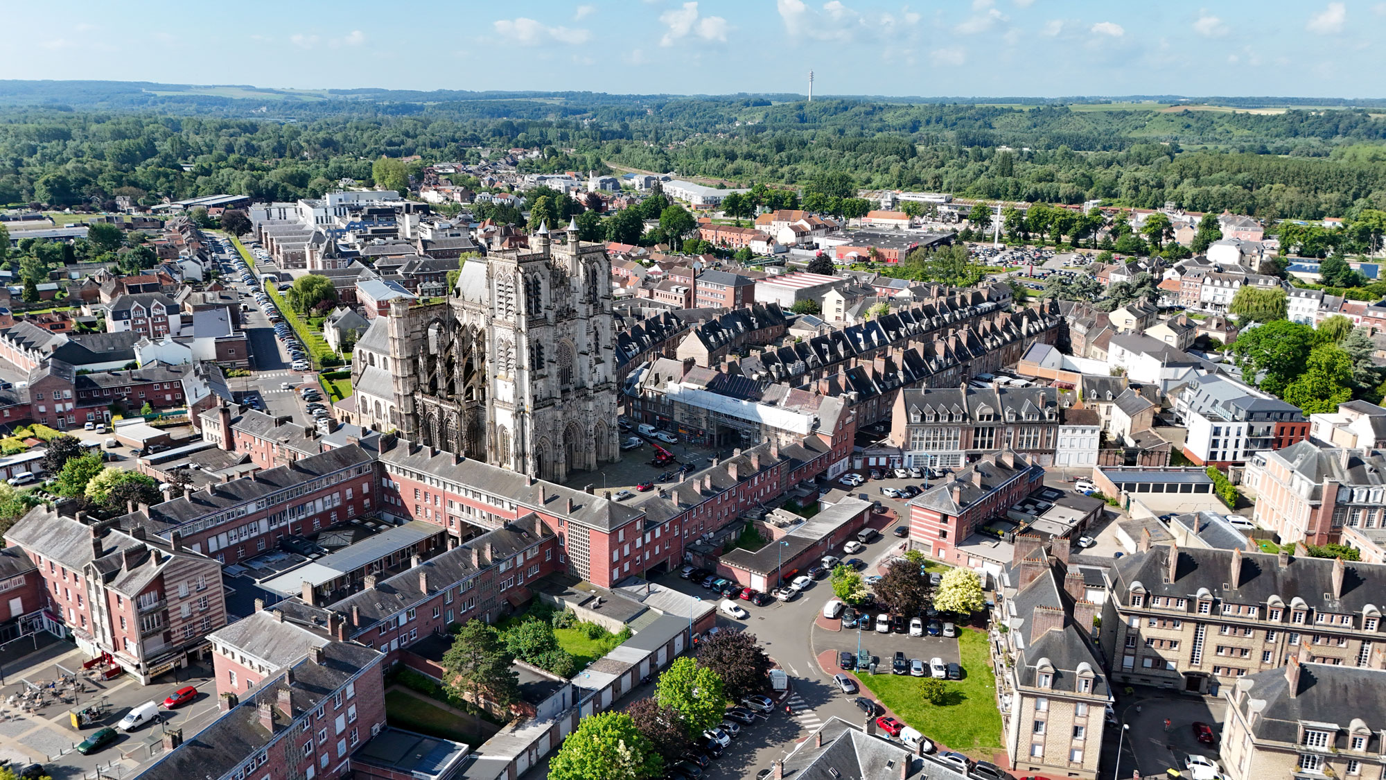 Vue aérienne d'une ville avec une cathédrale gothique imposante entourée de bâtiments historiques en briques. Les rues s'enroulent autour des structures et la verdure est visible au loin sous un ciel bleu parsemé de nuages.