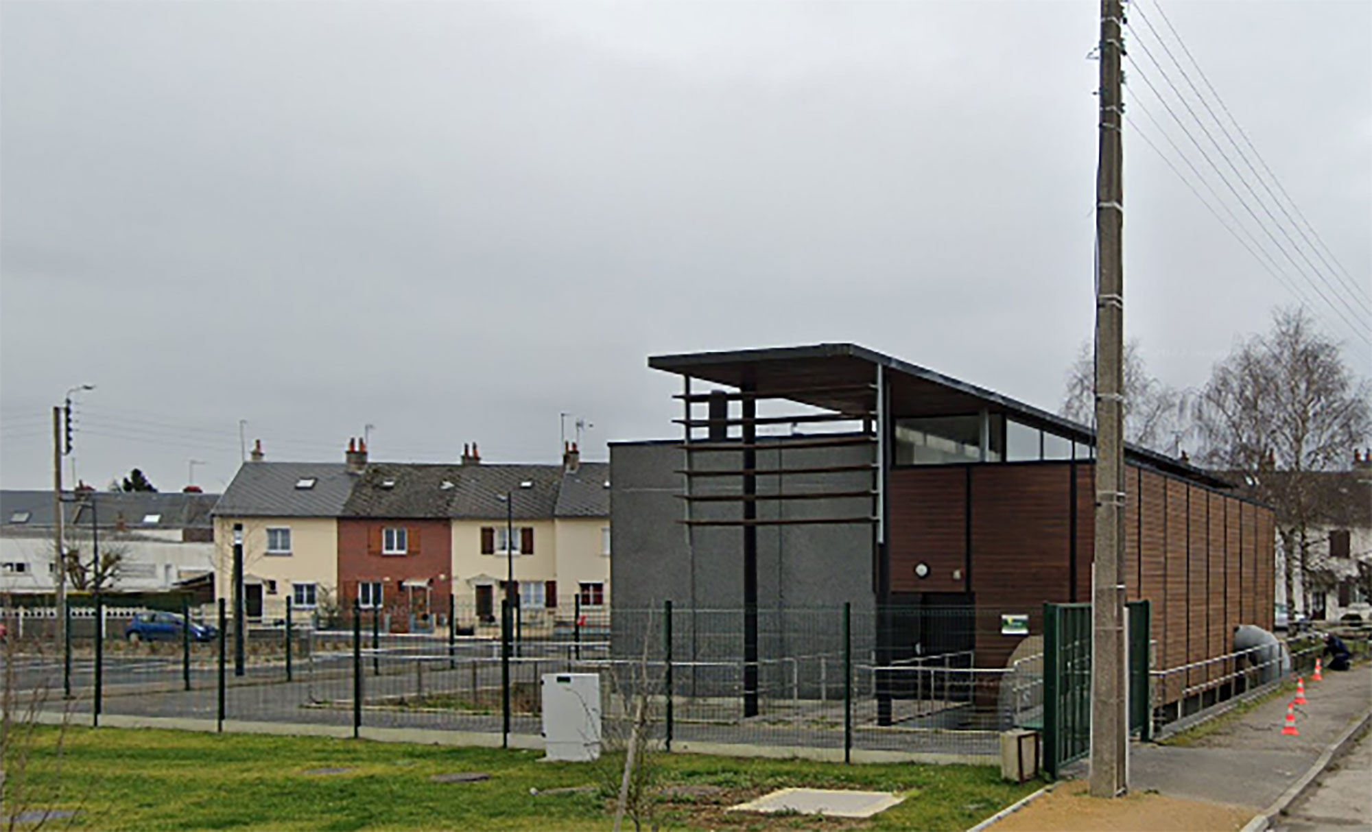 Un bâtiment moderne avec un extérieur en bois et en béton est entouré d'une clôture métallique. À l'arrière-plan, on voit des maisons résidentielles à deux étages avec des cheminées et un ciel nuageux au-dessus. Un poteau électrique et une zone herbeuse sont au premier plan.