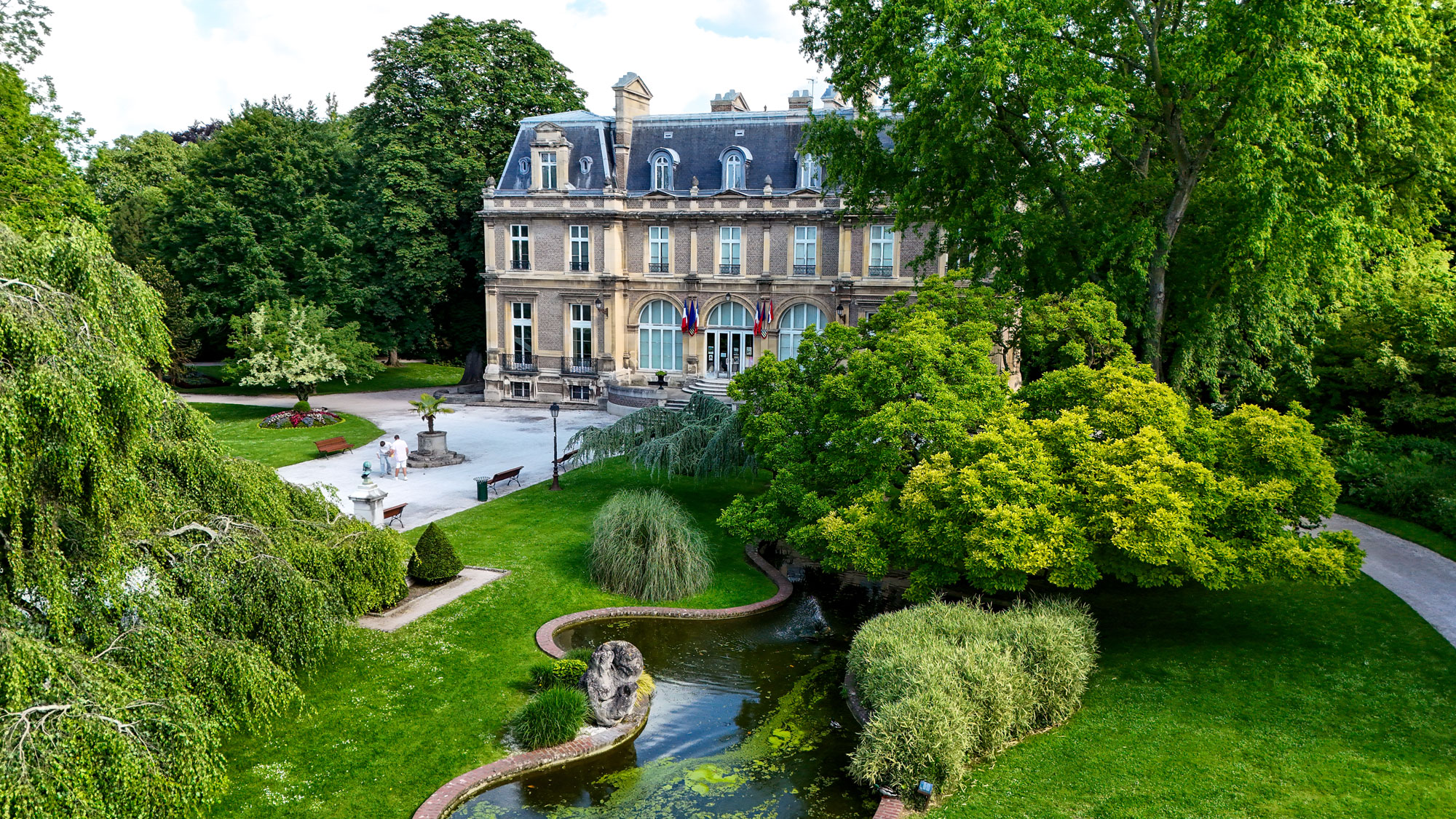 Un grand château au toit d'ardoise et aux détails architecturaux classiques se dresse au milieu d'une végétation luxuriante. Un jardin bien entretenu comprend un étang avec une petite fontaine, entouré d'arbres et de sentiers, créant un paysage serein et pittoresque.