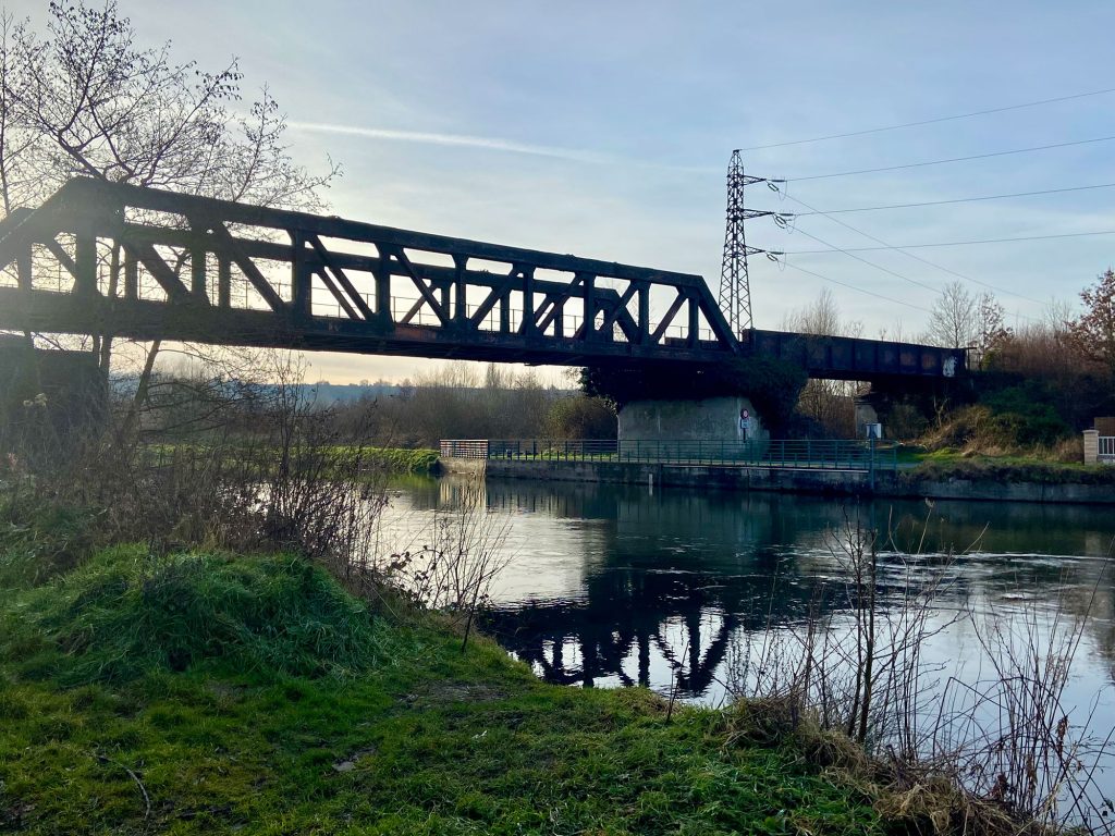 Un pont ferroviaire métallique enjambe une rivière calme. Les environs sont composés d'herbe verte et d'arbres nus, avec un ciel bleu clair au-dessus. Des lignes électriques sont visibles en arrière-plan.