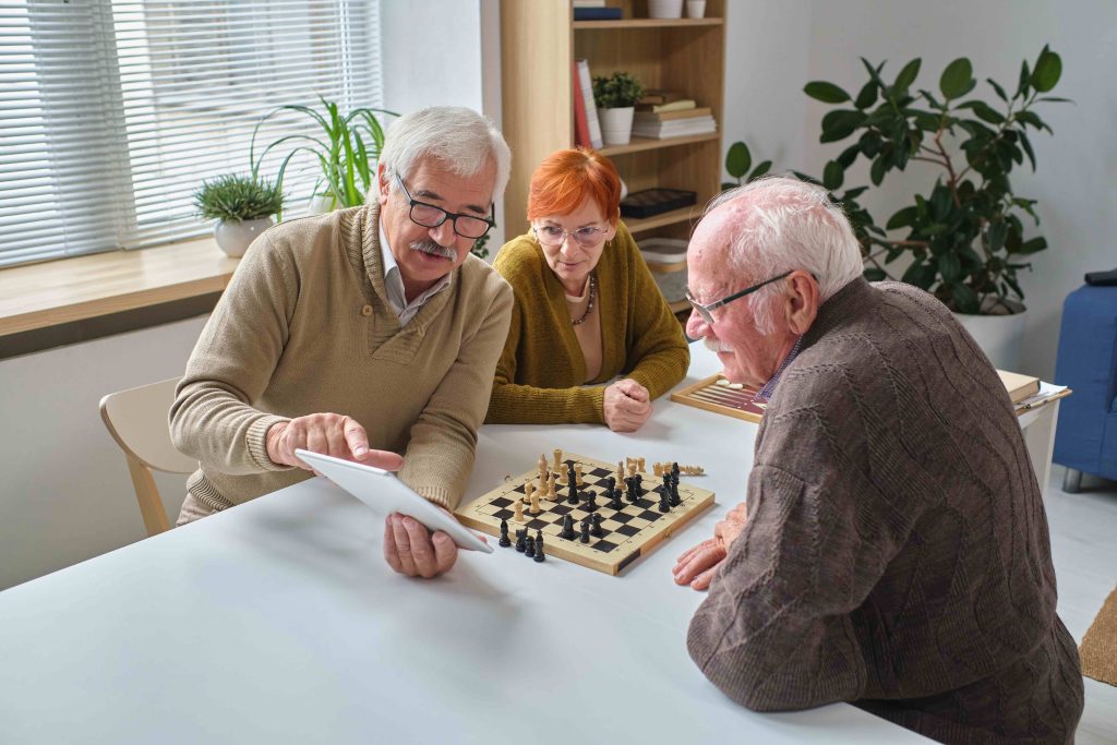 Trois personnes âgées sont assises à une table avec un échiquier. L'un d'eux pointe du doigt l'écran d'une tablette tandis que les deux autres écoutent attentivement. La pièce est bien éclairée avec des plantes et une bibliothèque en arrière-plan.