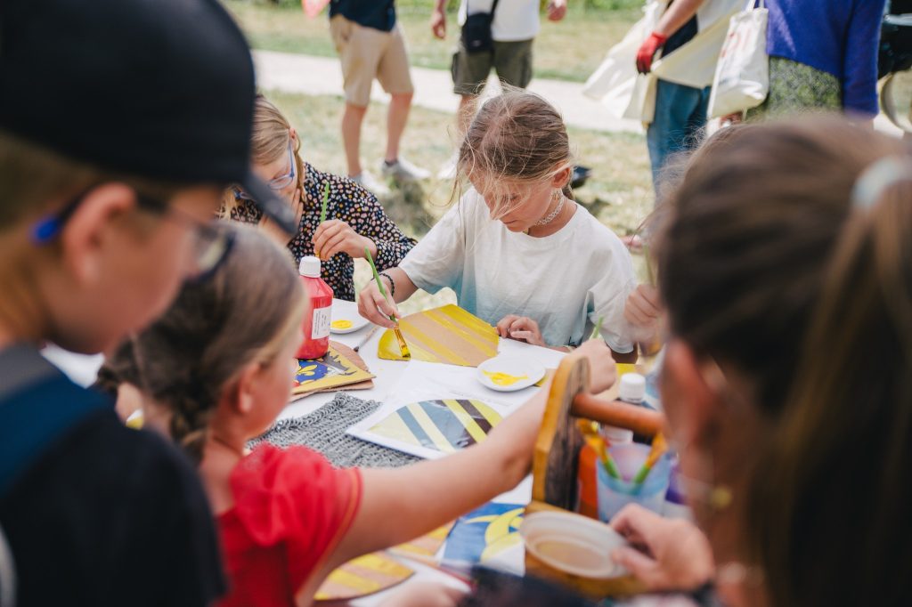 Un groupe d'enfants s'adonnent à des activités de peinture autour d'une table en plein air, entourés de fournitures artistiques. Un enfant se concentre sur une toile à rayures jaunes et noires. Ils semblent concentrés et apprécient le processus créatif.