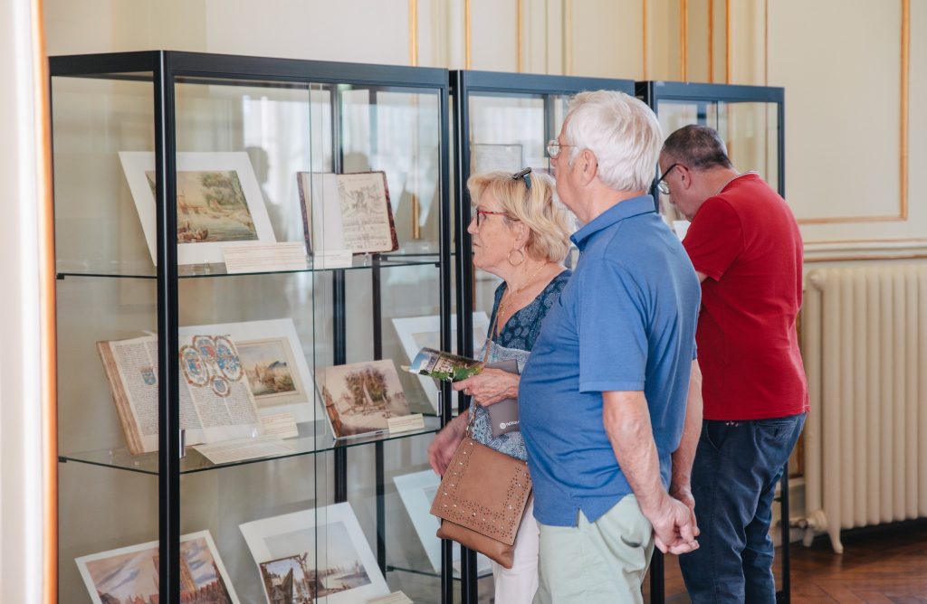 Un couple de personnes âgées examine des objets historiques dans une vitrine de musée, tandis qu'un homme en chemise rouge examine une autre exposition. La pièce est élégamment décorée de murs jaunes et de moulures décoratives.