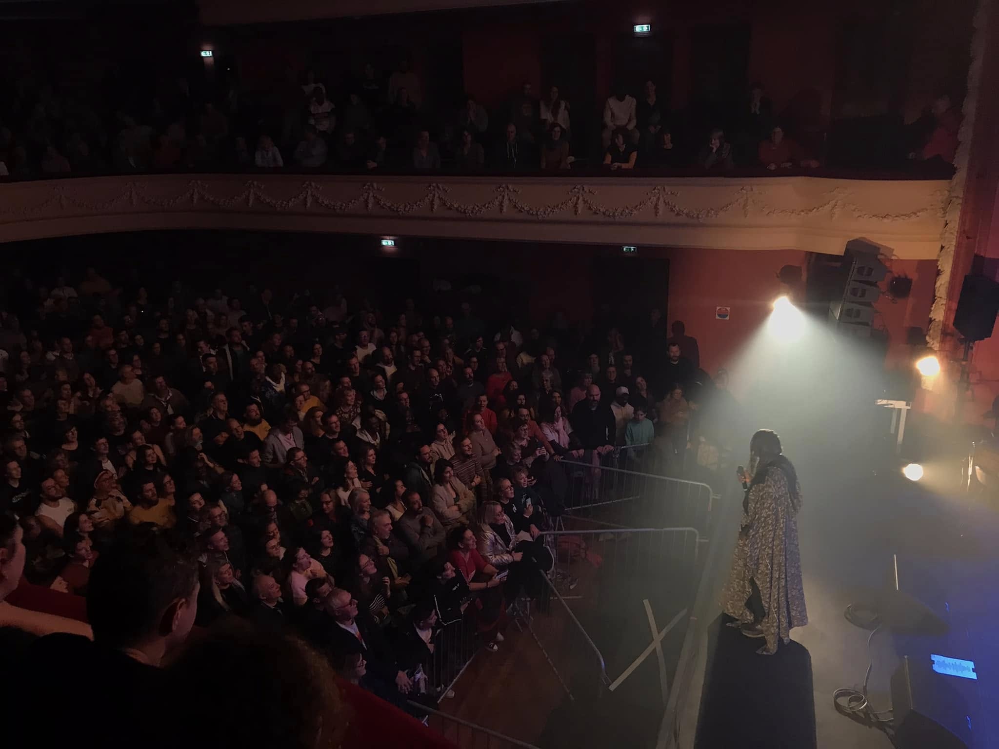 Un artiste sur scène, vêtu d'une robe, s'adresse à un public comble dans un théâtre faiblement éclairé. Le public regarde attentivement, assis sur plusieurs niveaux. L'atmosphère est intime et concentrée.