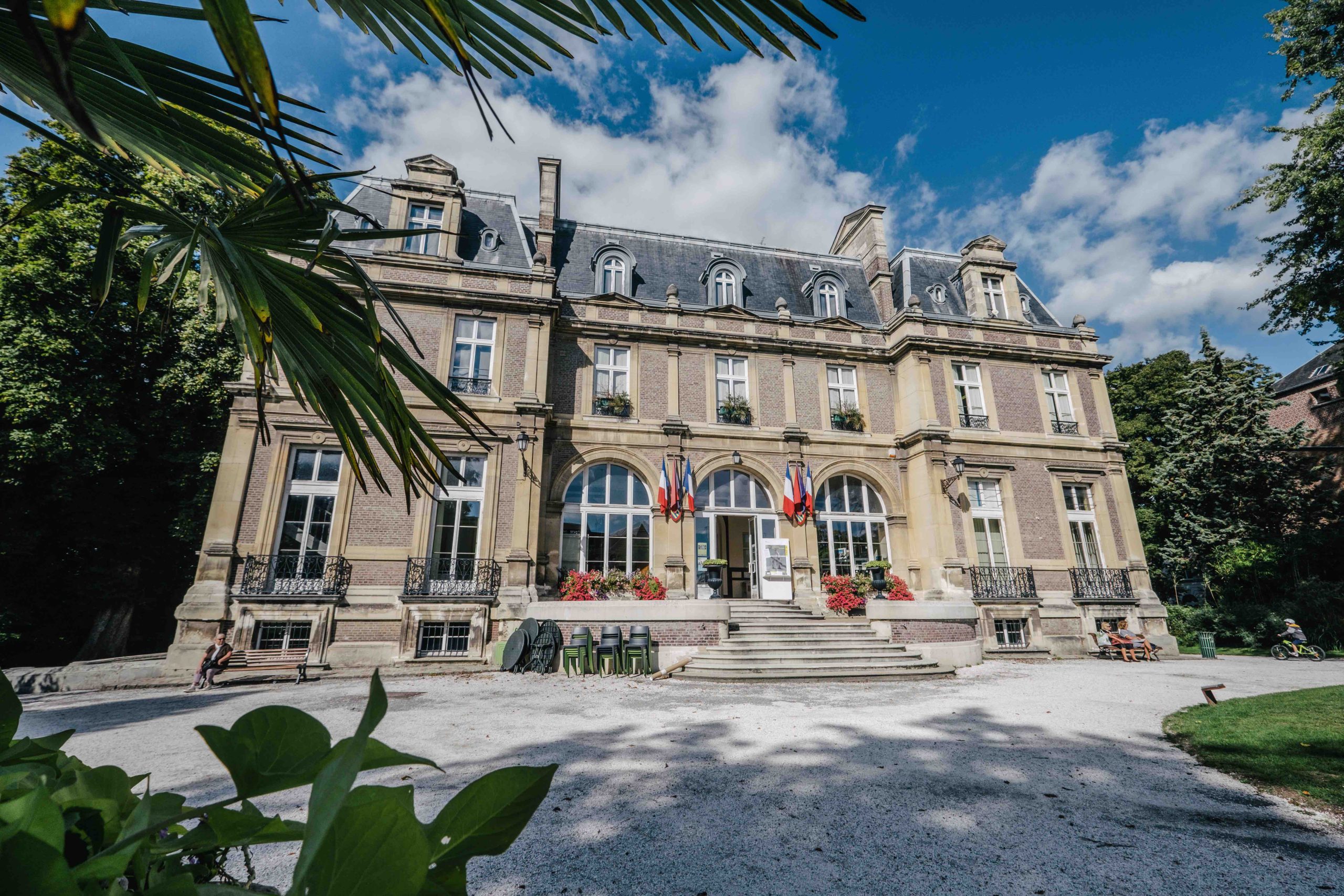 Un grand bâtiment historique avec une façade symétrique, de grandes fenêtres et un toit en pente sous un ciel bleu. Des drapeaux ornent l'entrée et une végétation luxuriante entoure la scène, encadrant magnifiquement la structure.