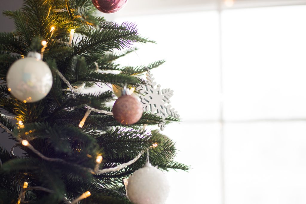 Un sapin de Noël décoré de boules blanches et roses, d'un flocon de neige et de guirlandes lumineuses. Une douce lumière naturelle filtre à travers une fenêtre en arrière-plan.