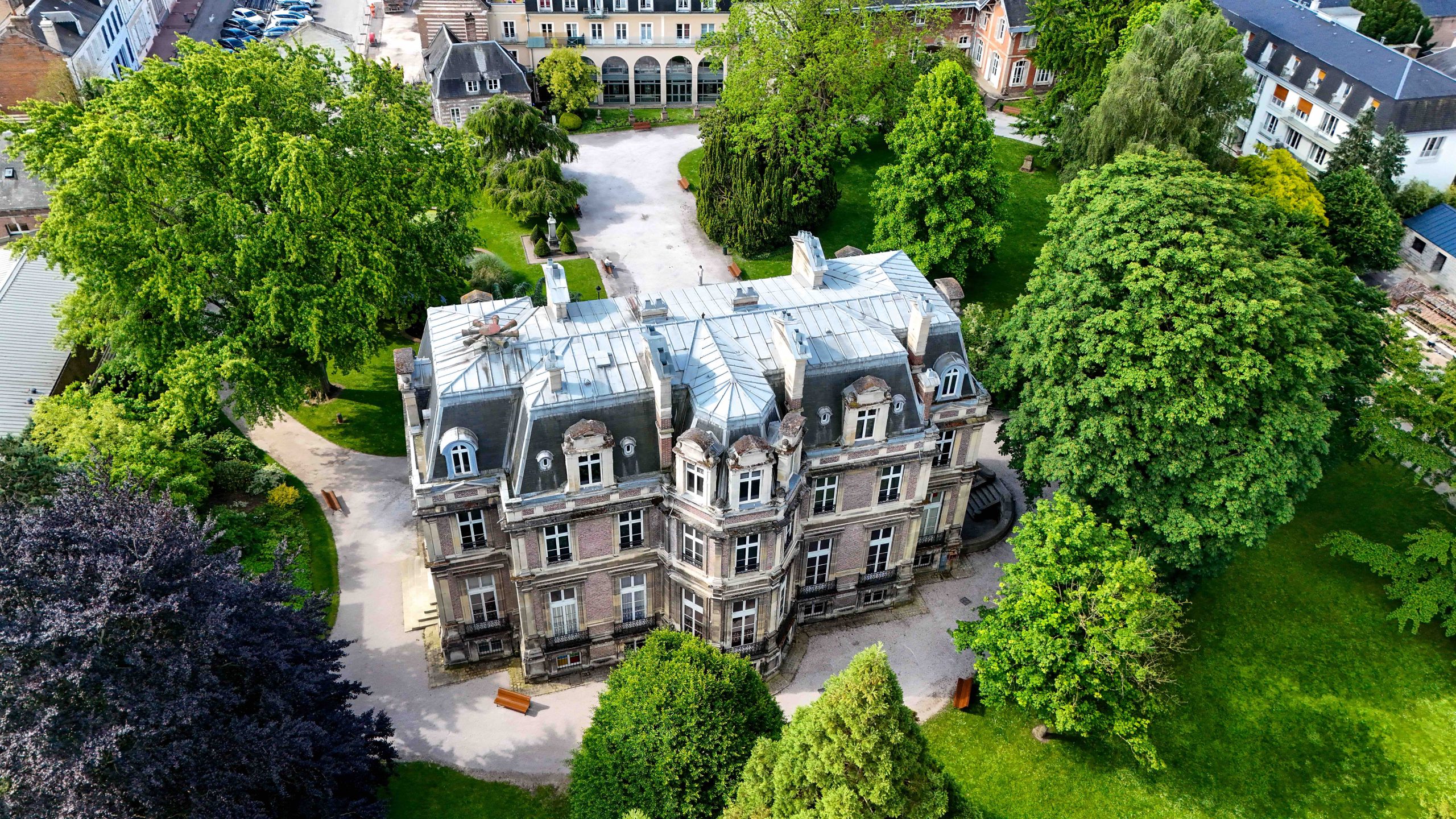 Vue aérienne d'un bâtiment historique de trois étages entouré d'arbres et de jardins verdoyants. L'architecture présente un toit mansardé et une façade en pierre. Des sentiers entourent le bâtiment, niché dans un cadre serein et paysager.