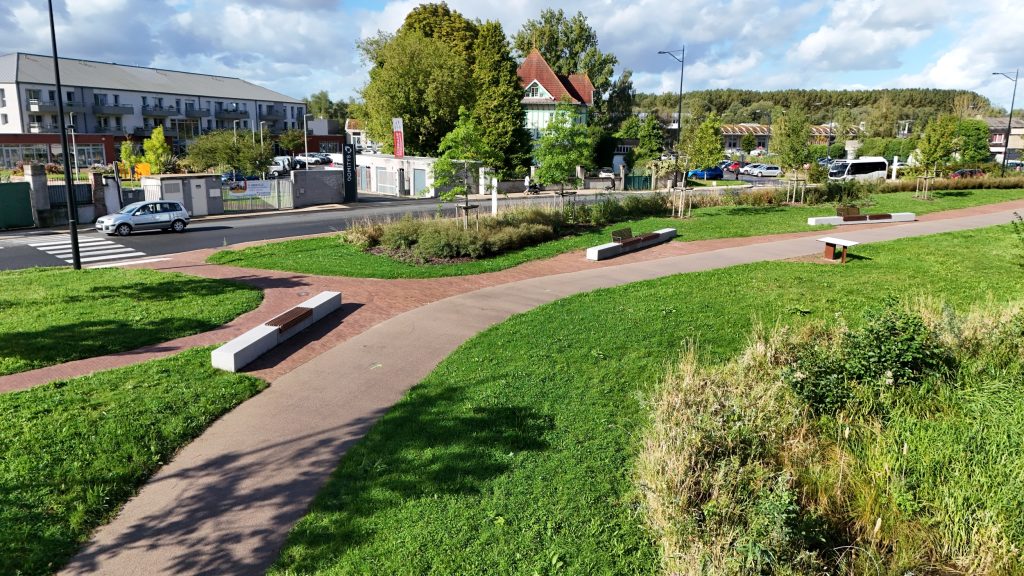 Un parc avec des sentiers courbes, des bancs et des espaces verts bien entretenus. Des arbres et des arbustes bordent la zone. Des bâtiments et une rue avec un passage piéton sont visibles en arrière-plan sous un ciel partiellement nuageux.