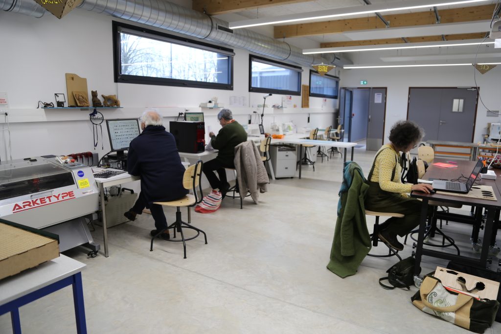 Un atelier moderne avec trois personnes travaillant à des bureaux séparés. Une personne utilise un ordinateur à proximité d'une machine de marque « Arketype ». La salle dispose de grandes fenêtres, d'un éclairage lumineux et de divers équipements techniques sur les tables.